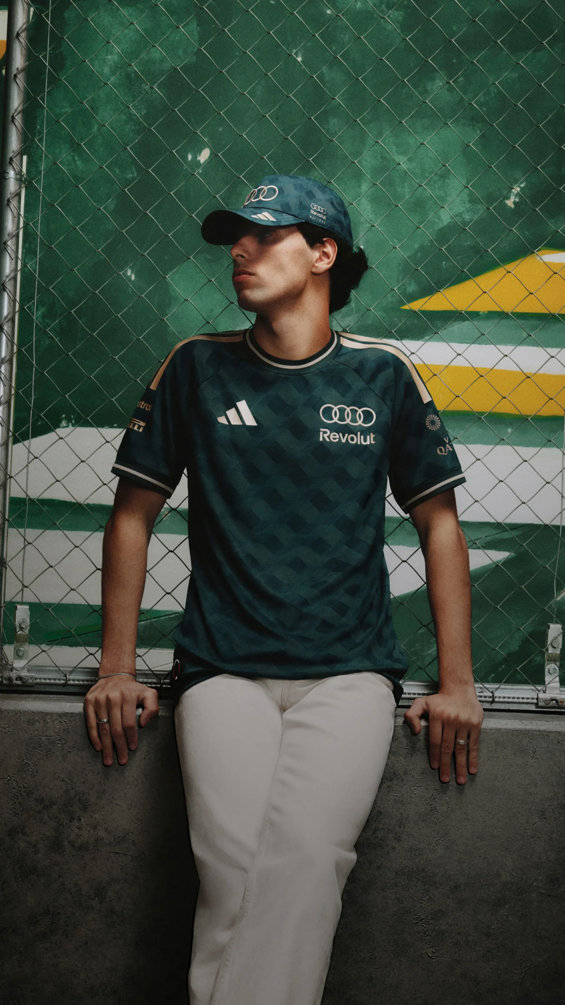 Audi Revolut F1® Team driver Gabriel Bortoleto in São Paulo jersey and cap leaning against a fence by a green and yellow wall