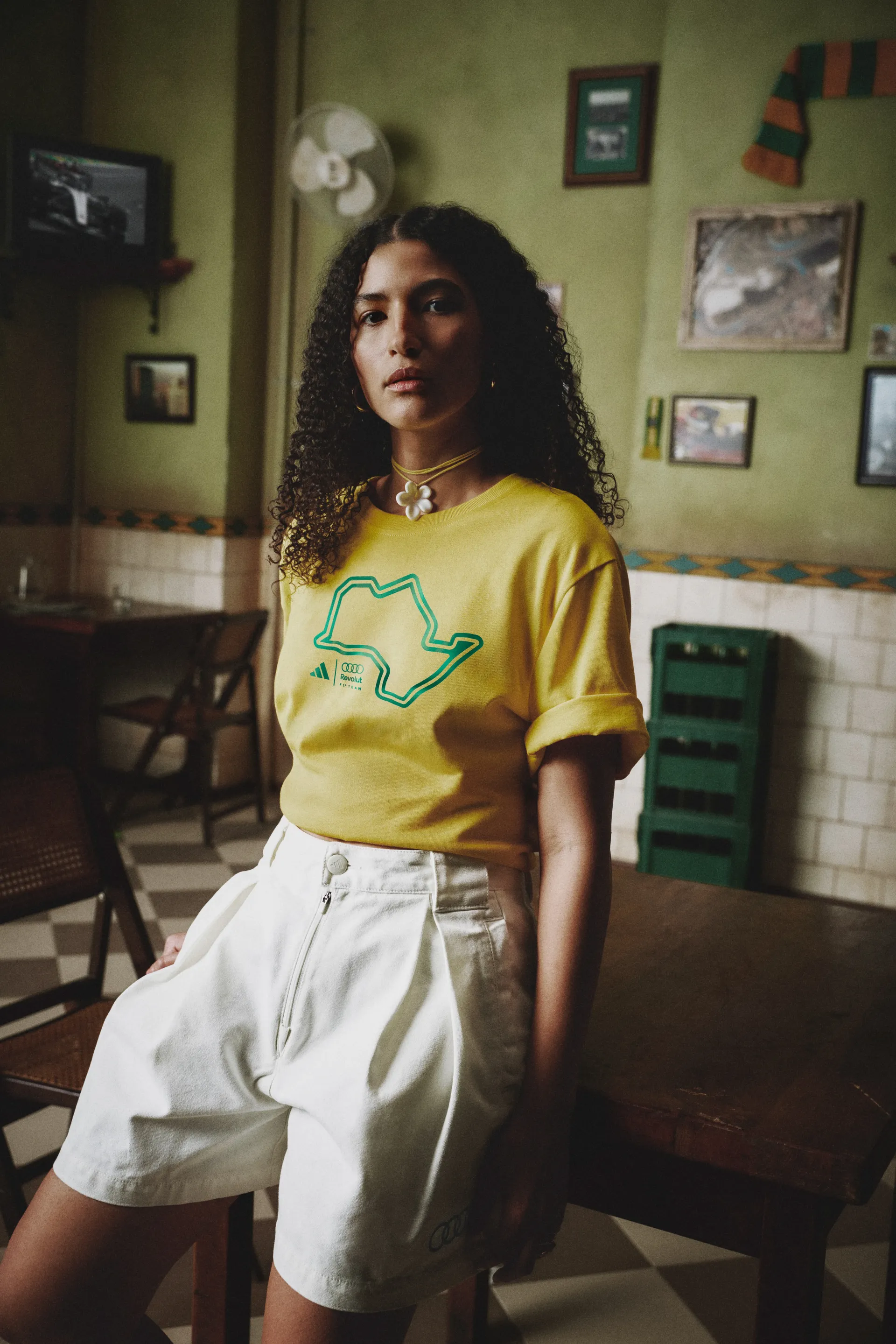 Model in a yellow graphic T-shirt and white shorts in a café, adidas x Audi Revolut F1® Team Sampa Soul collection