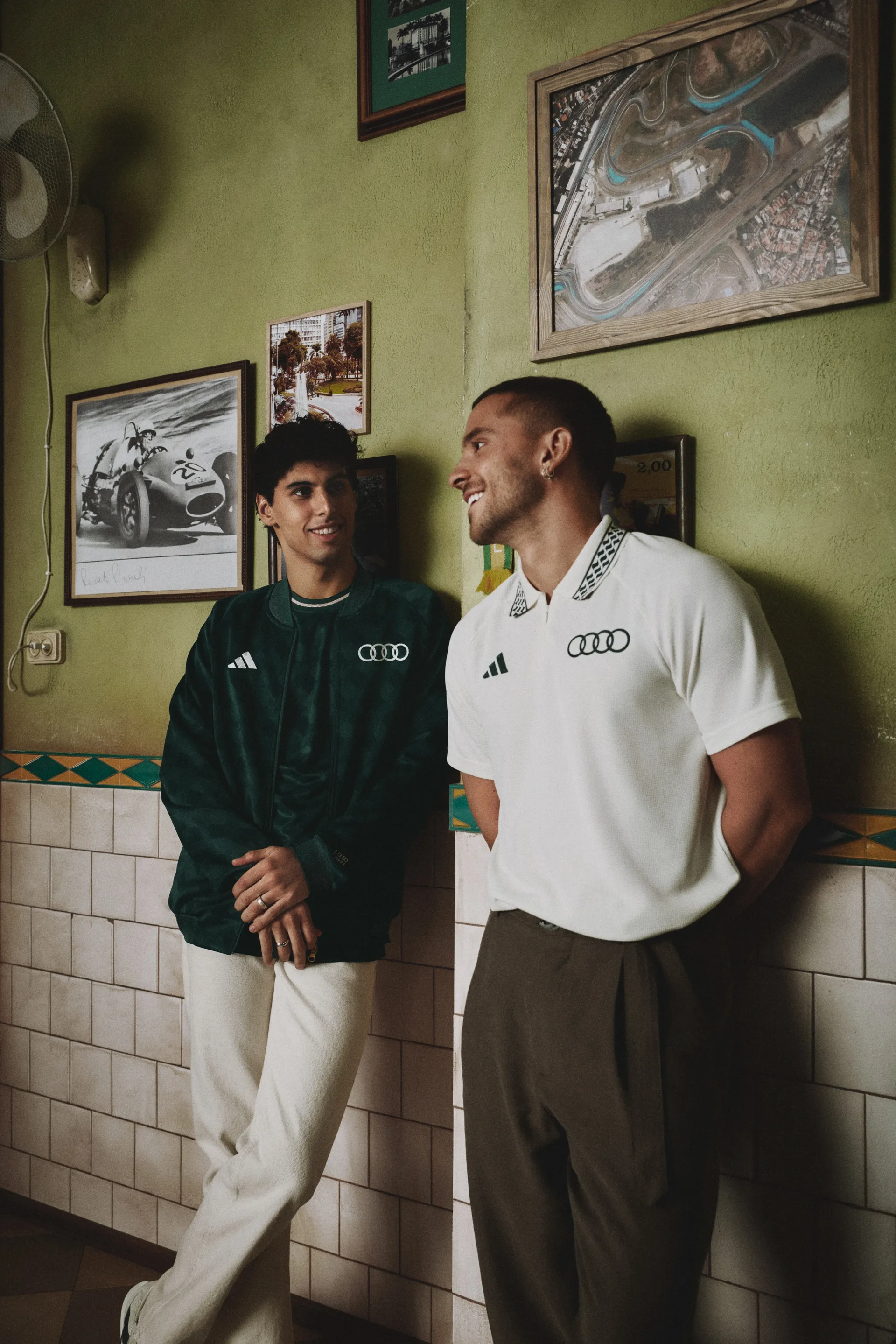Audi Revolut F1® Team driver Gabriel Bortoleto and male model leaning on a wall in a green track jacket and white polo, adidas x Audi Revolut F1® Team Sampa Soul collection