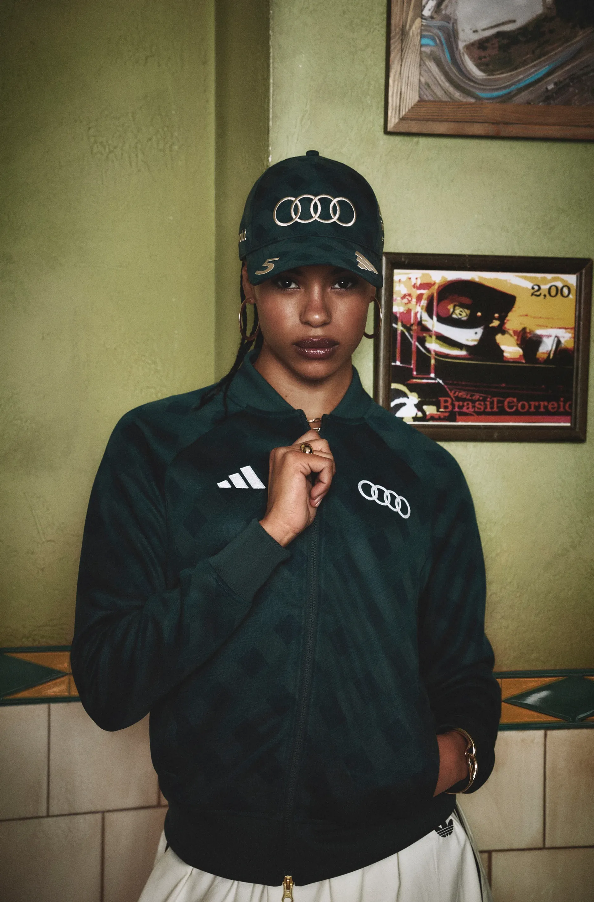 Model zipping up the adidas x Audi Revolut F1® Team São Paulo track jacket, wearing a matching cap indoors