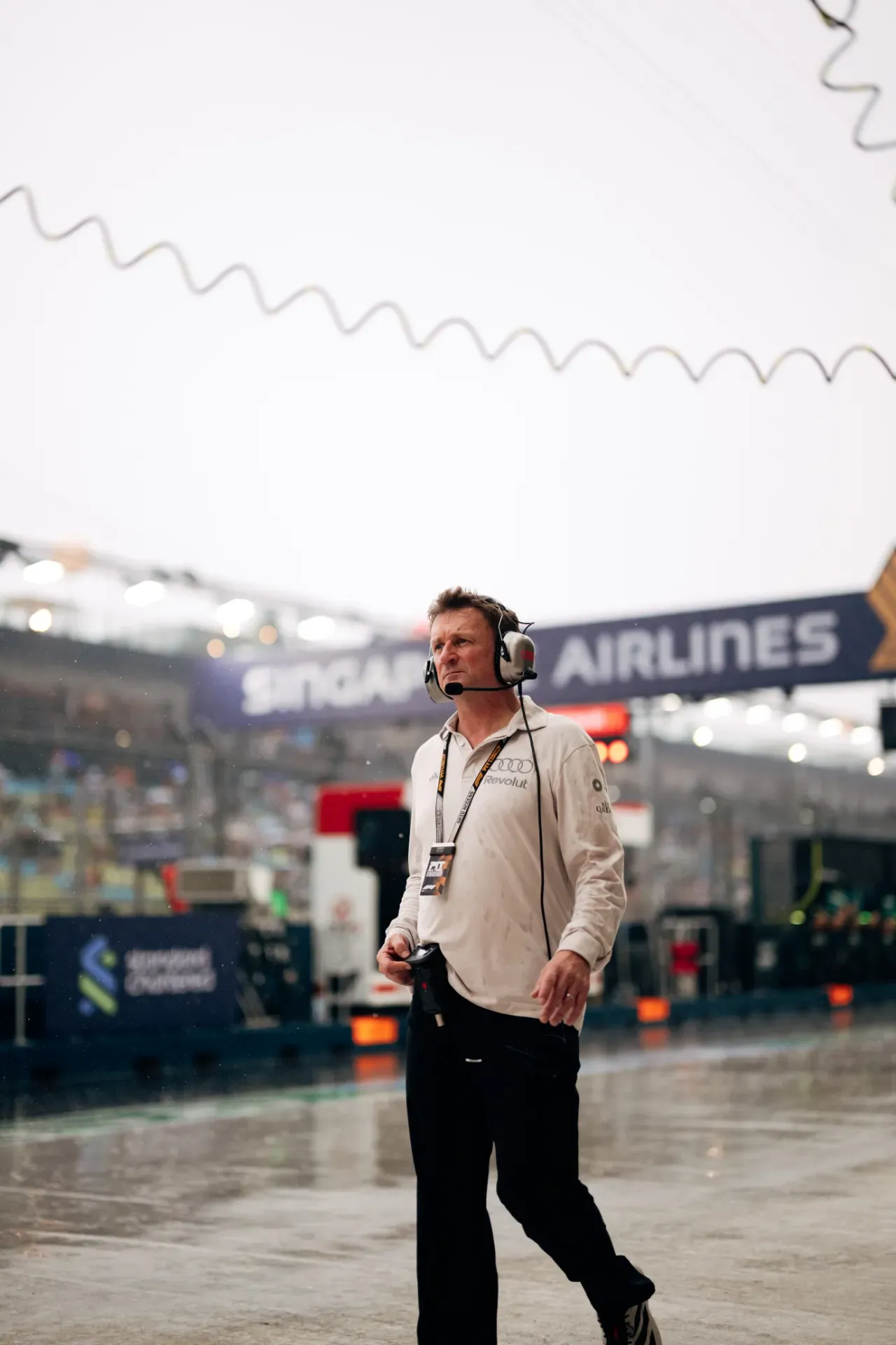 Allan McNish of the Audi Revolut F1® Team wearing a headset and lanyard walking along the wet pit lane in Singapore