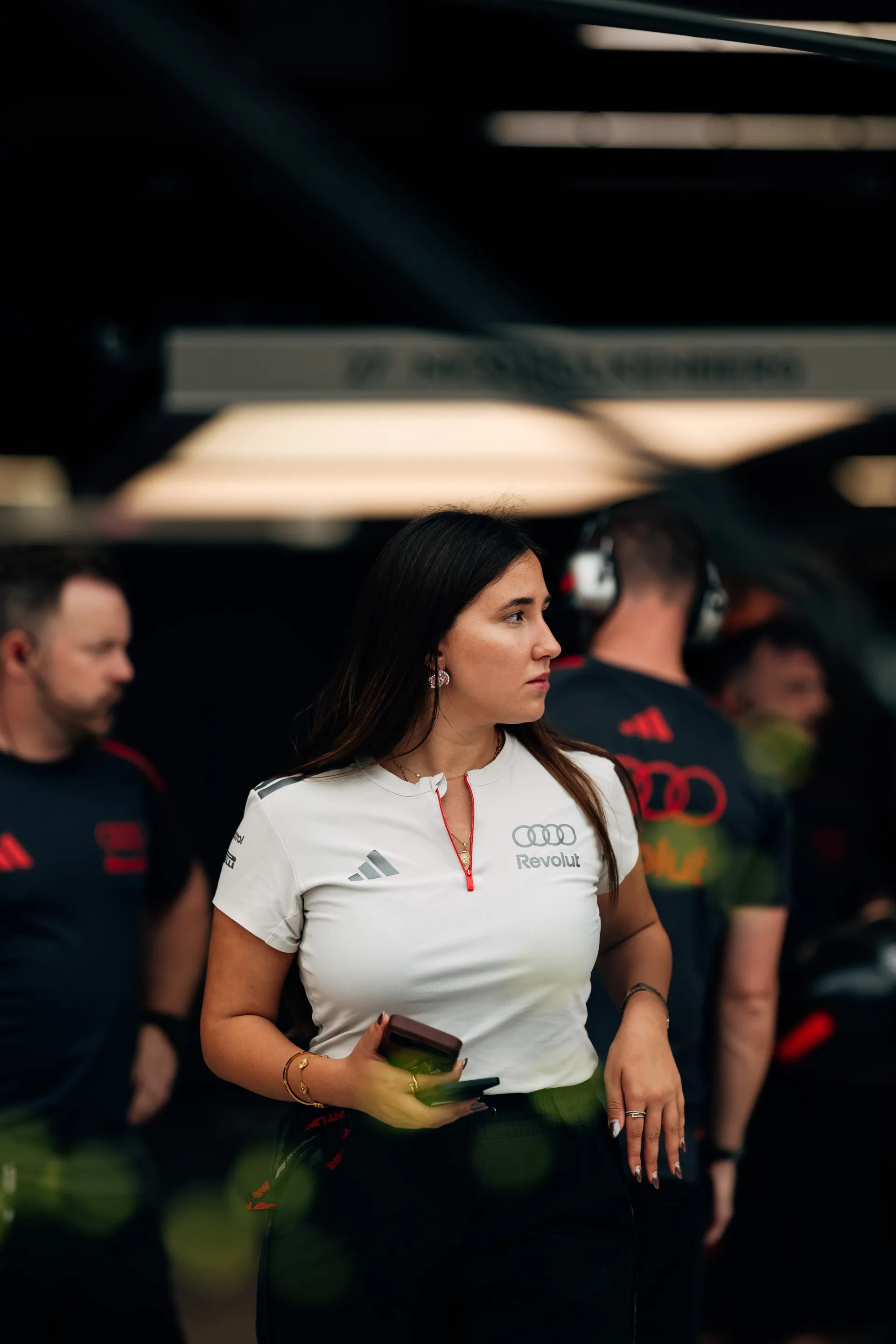 Audi Revolut F1® Team engineer in a white team shirt holding her phone in the garage, mechanics working behind her.