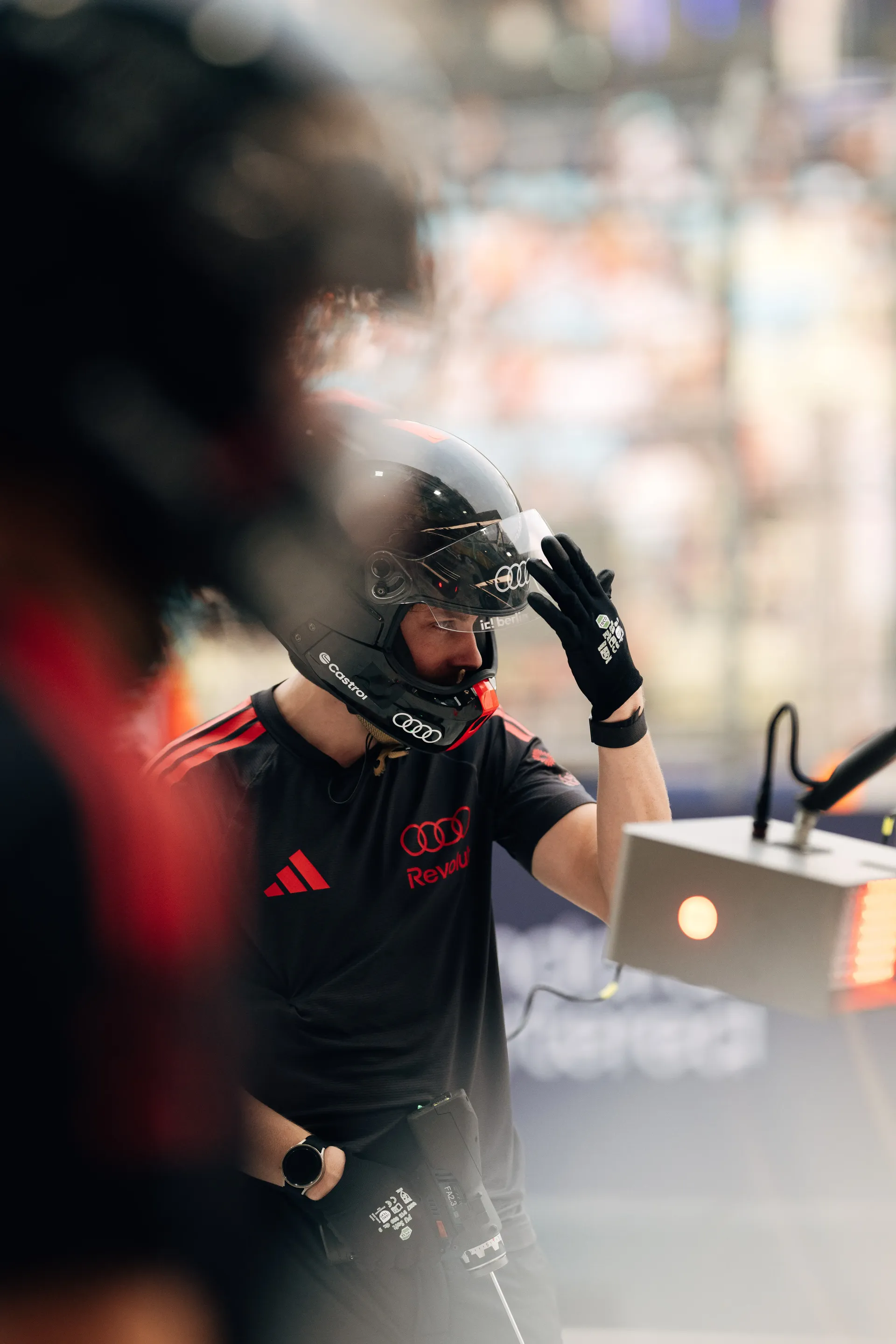 Audi Revolut F1® Team mechanic in the pit lane, framed by a crew member blurred in the foreground.