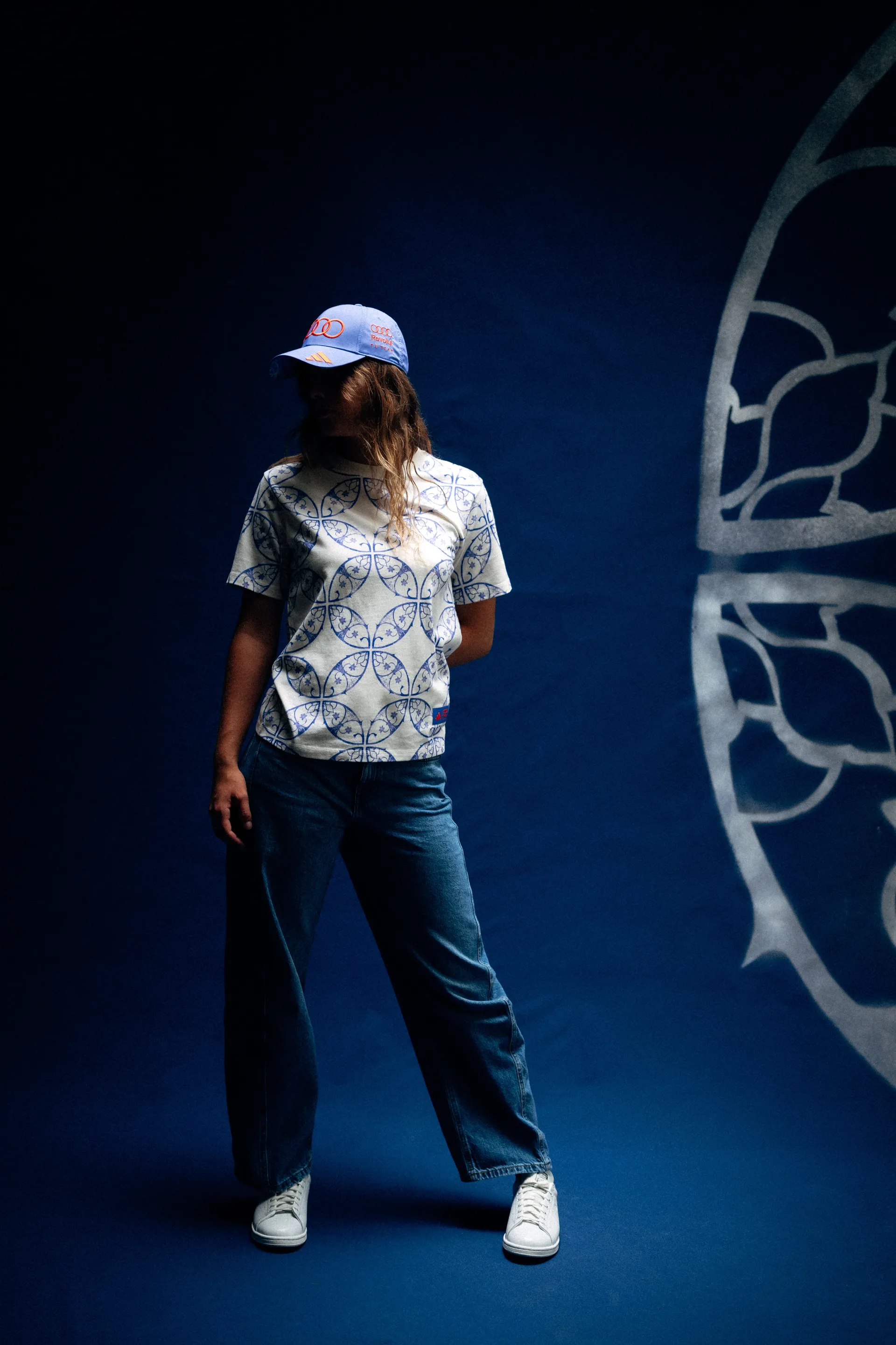 Model im T-Shirt des Audi Revolut F1® Teams mit durchgehendem Kachelprint, Cap, Jeans und Sneakers