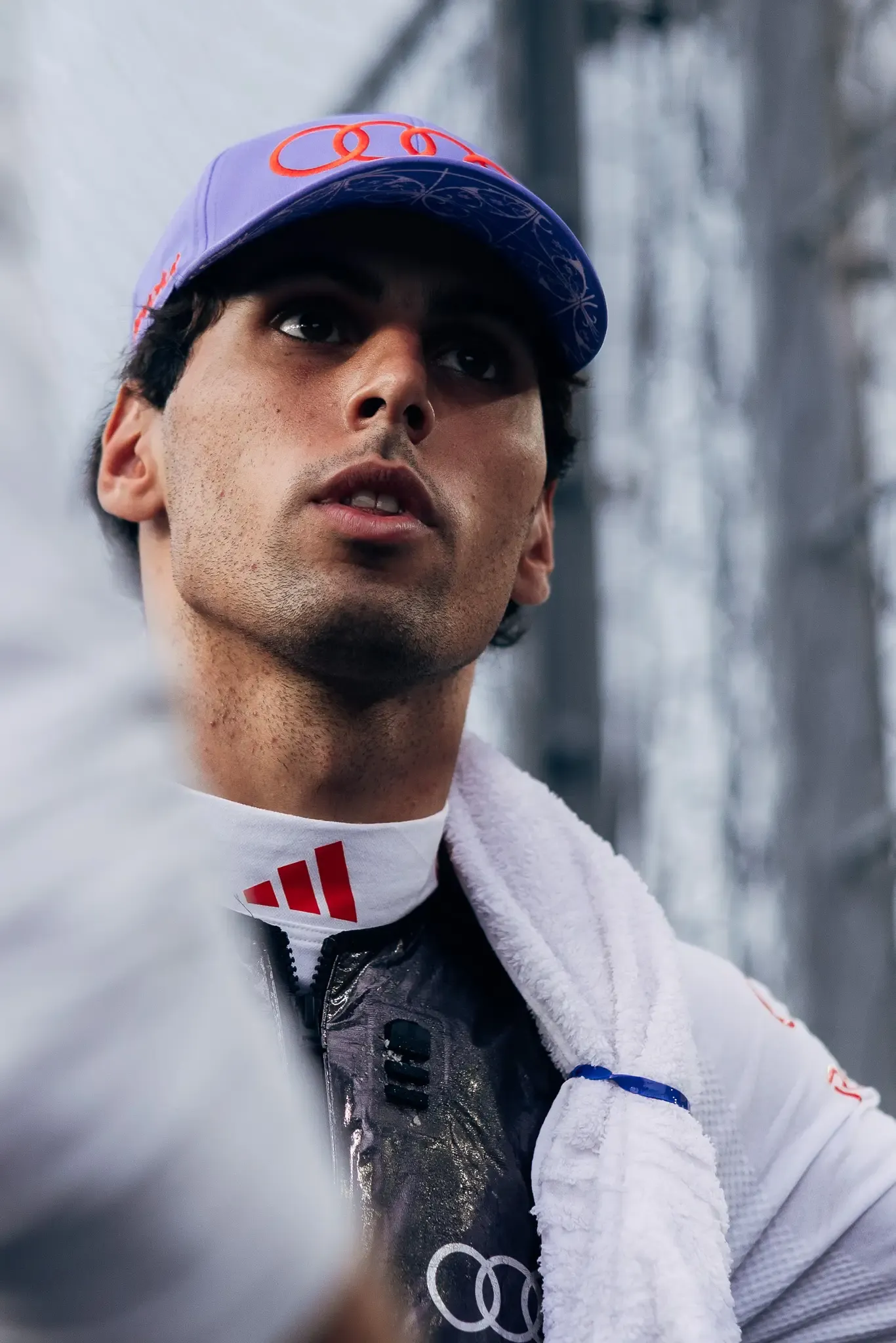 Audi Revolut F1® Team driver Gabriel Bortoleto in a lilac cap and white team kit with a towel around his neck, looking up and off-frame in the Singapore paddock.