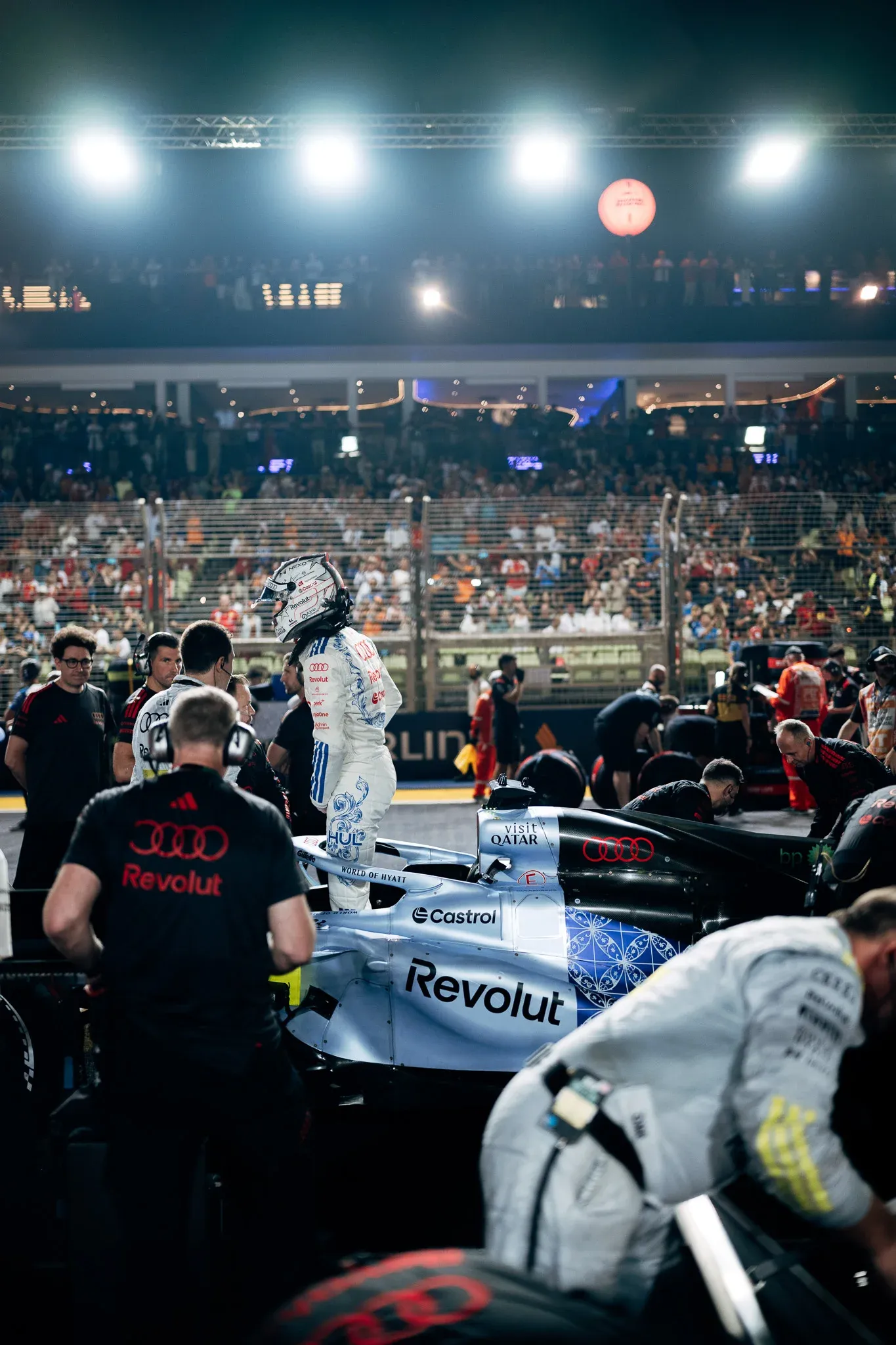 Audi Revolut F1® Team car R26 on the Singapore grid at night, surrounded by mechanics and engineers, with floodlights and packed grandstands behind.