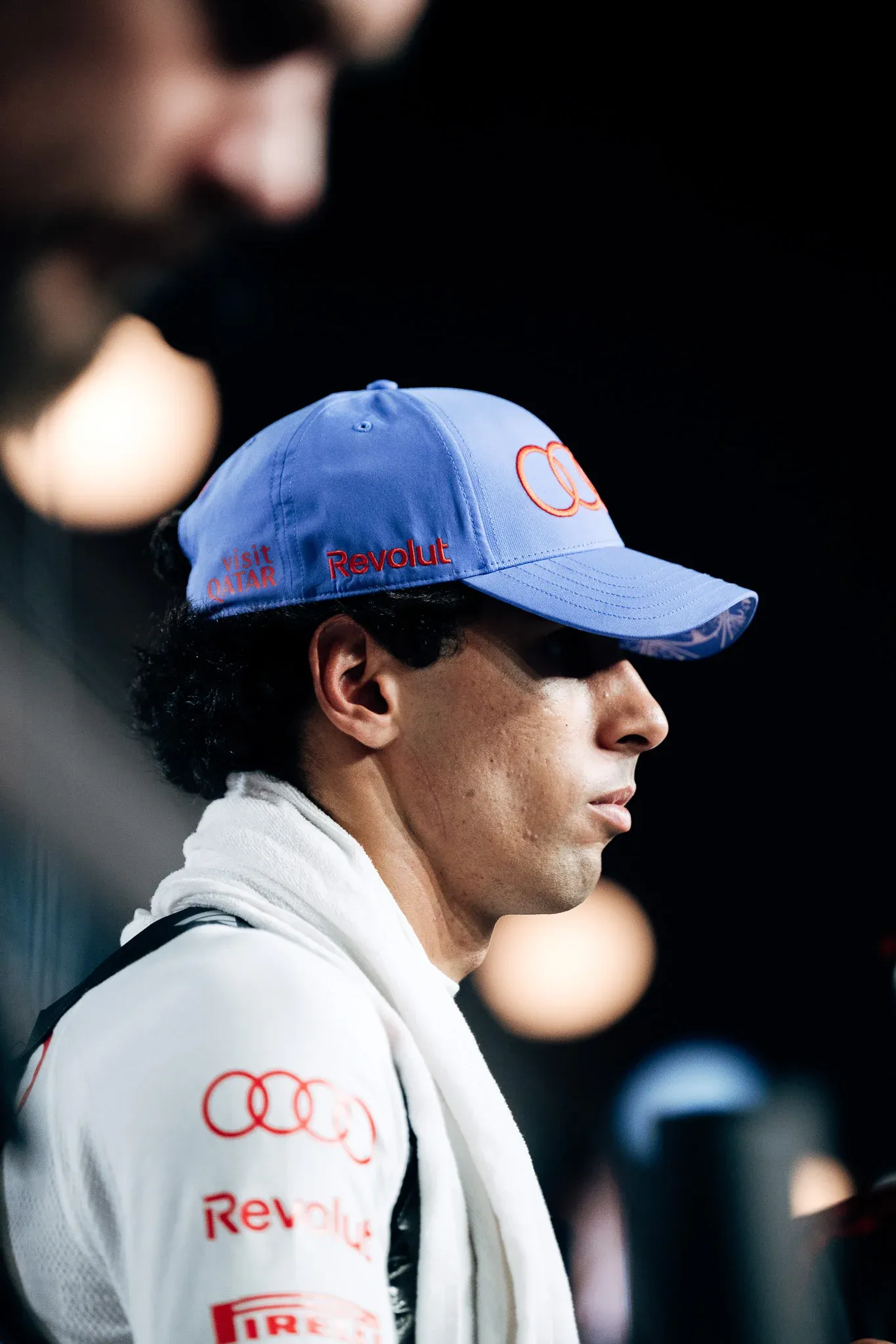 Audi Revolut F1® Team driver Gabriel Bortoleto in profile, wearing a blue cap with a towel around his neck, against a dark background.