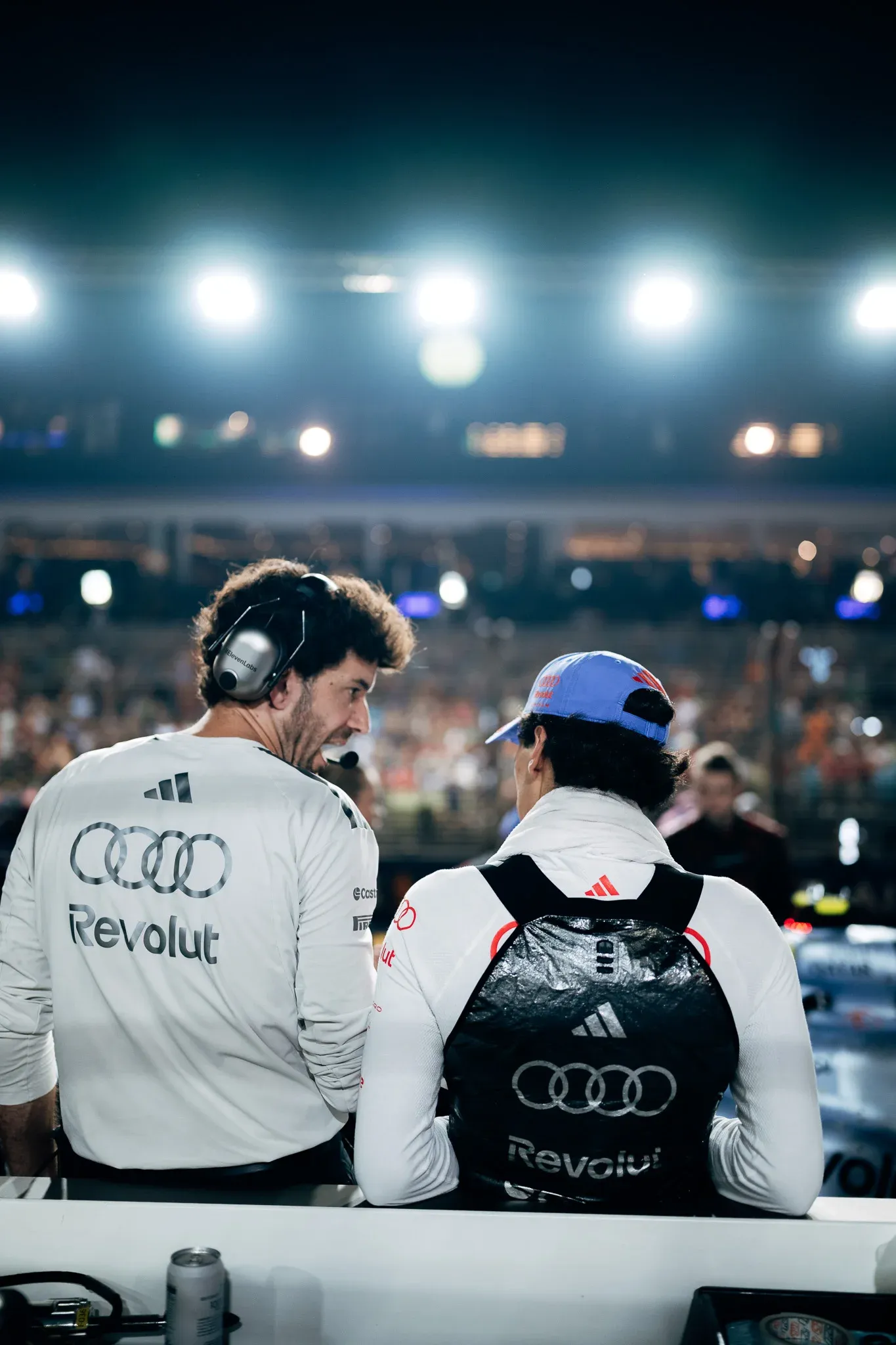 Audi Revolut F1® Team engineer in a white shirt and headset, seen from behind with Audi Revolut F1® Team driver Gabriel Bortoleto in a blue cap, facing the floodlit grandstands.