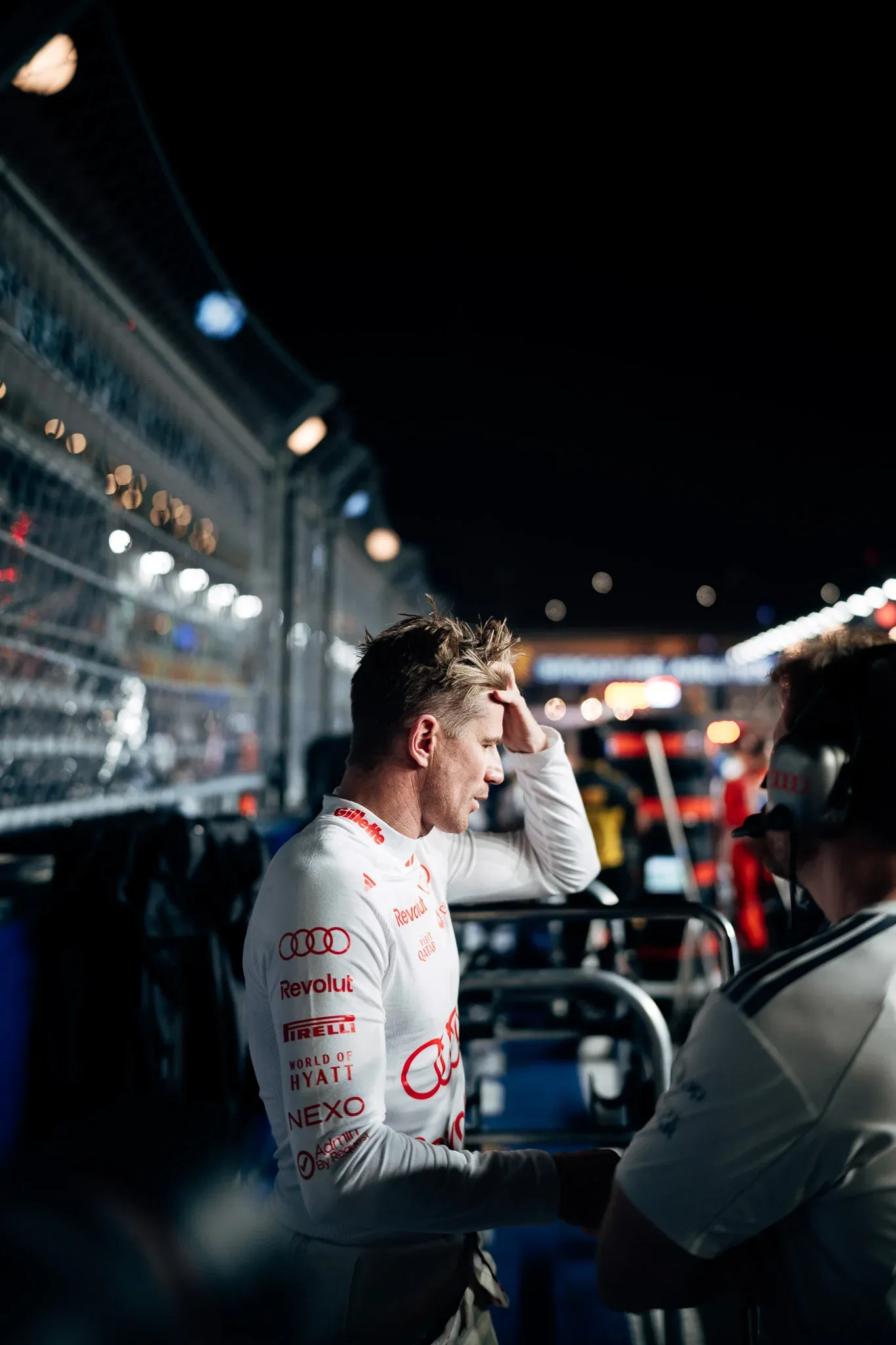 Audi Revolut F1® Team driver Nico Hulkenberg stands in the pit lane at night with a hand to his forehead, next to the catch fence.