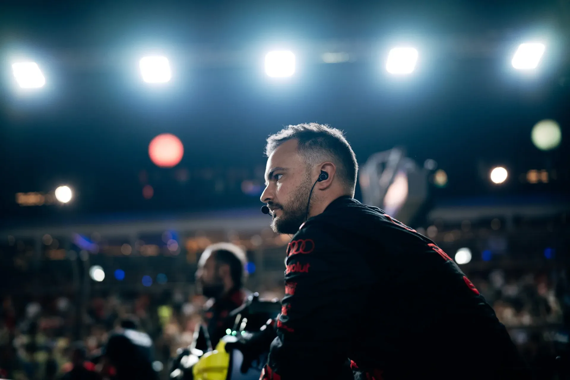 Audi Revolut F1® Team mechanic stands on the grid under bright floodlights, with the crowd blurred behind.