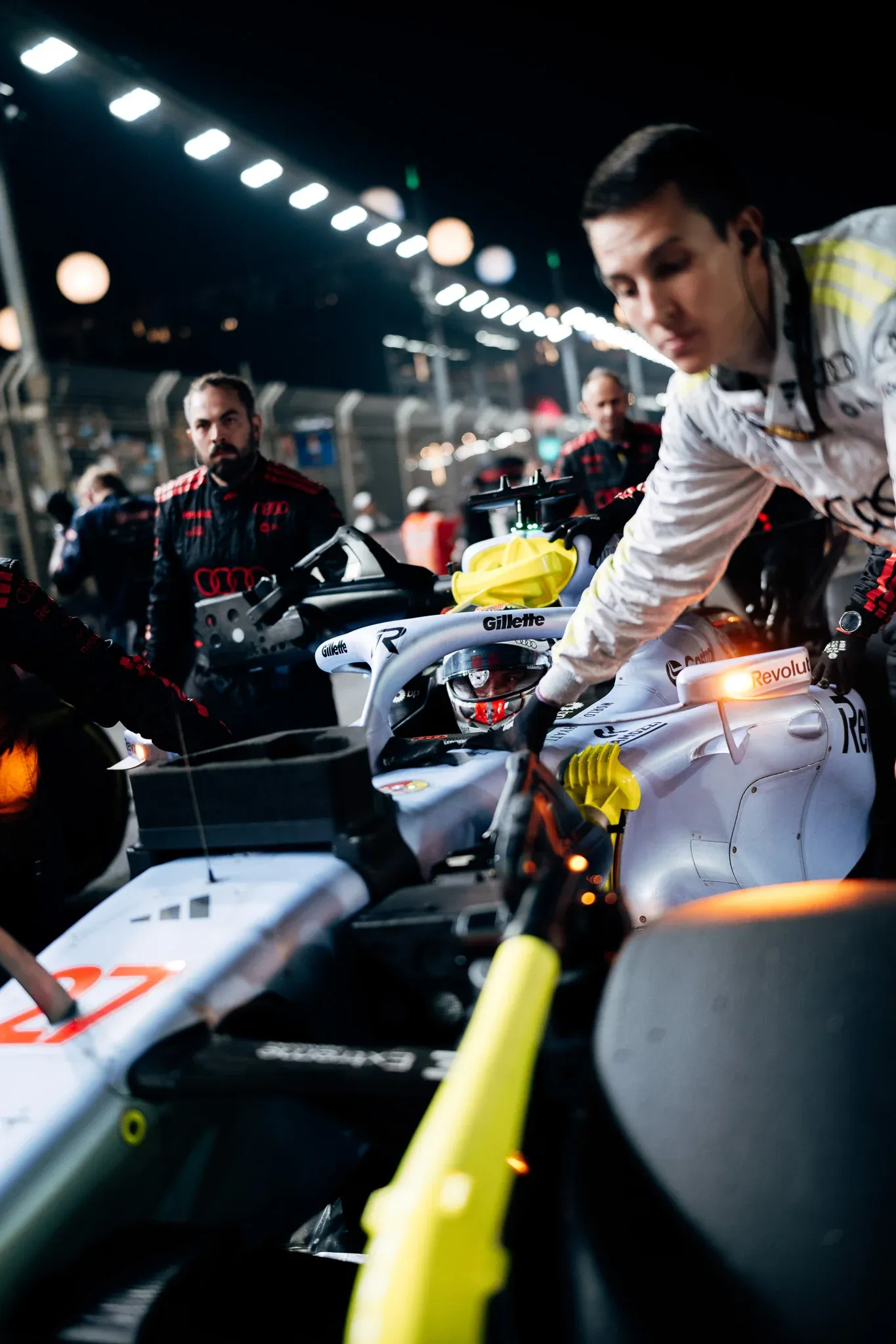 Audi Revolut F1® Team engineer leans over the cockpit of the car on the night grid as mechanics prepare it for the race.