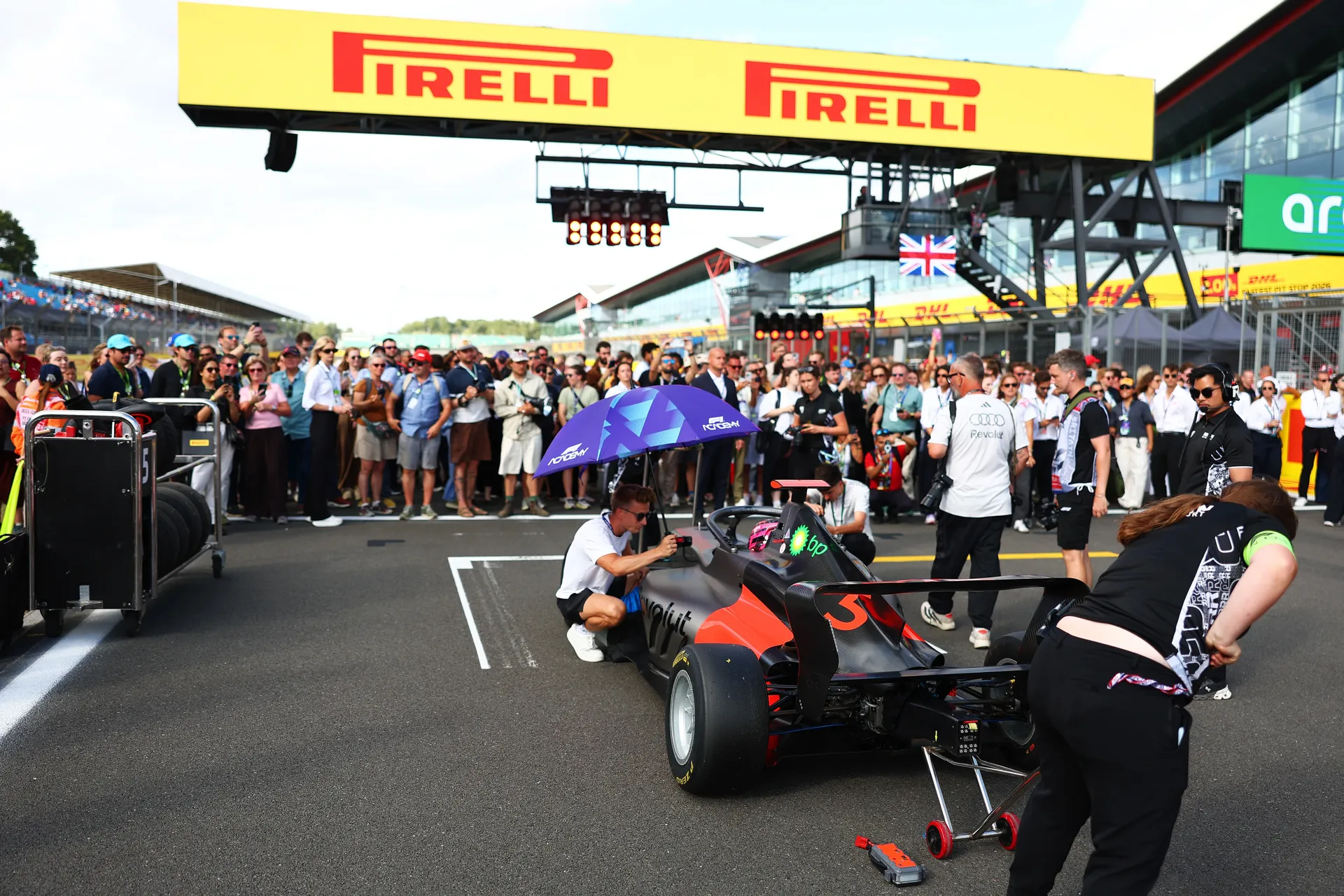 Audi Revolut F1® Team car is positioned on the Silverstone grid under a Pirelli gantry while crew members prepare the cockpit and fans gather behind barriers.