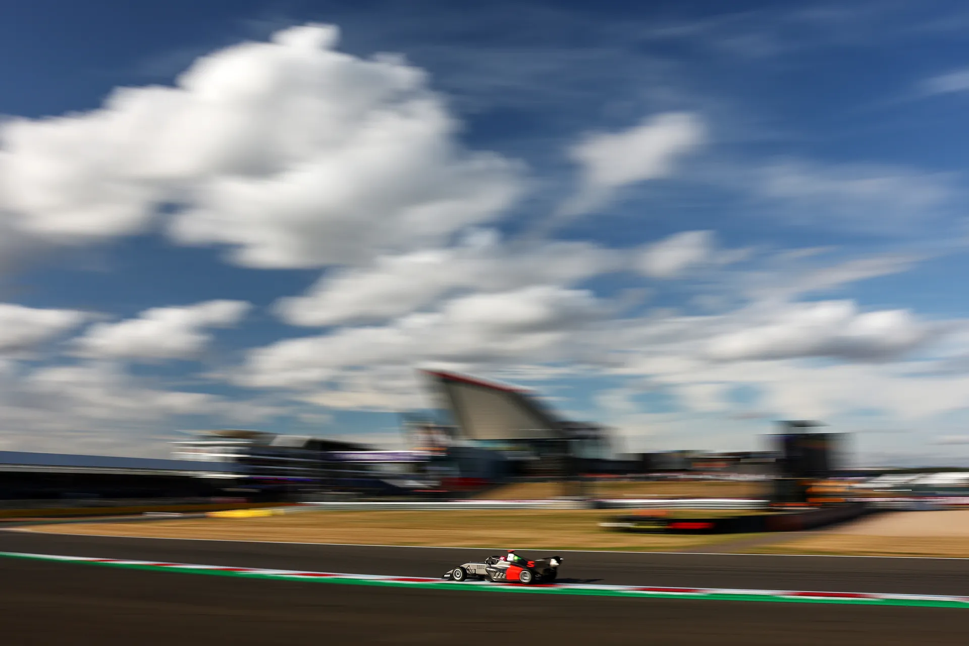 Audi Revolut F1® Team F1 Academy car #5 speeds along the Silverstone circuit with dramatic motion blur and wide sky above.