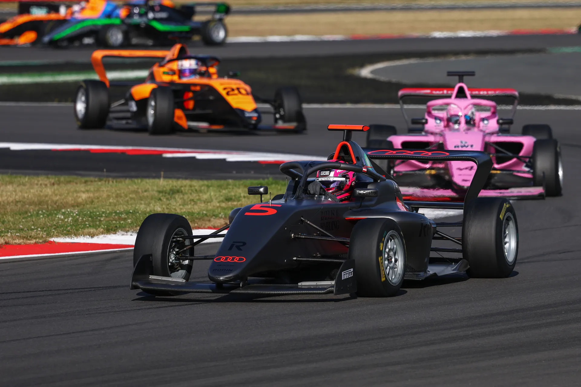 Emma Felbermayr drives the Audi Revolut F1® Team F1 Academy car #5 through a right-hand corner at Silverstone, leading ahead of a pink and an orange car during a close on-track race battle.