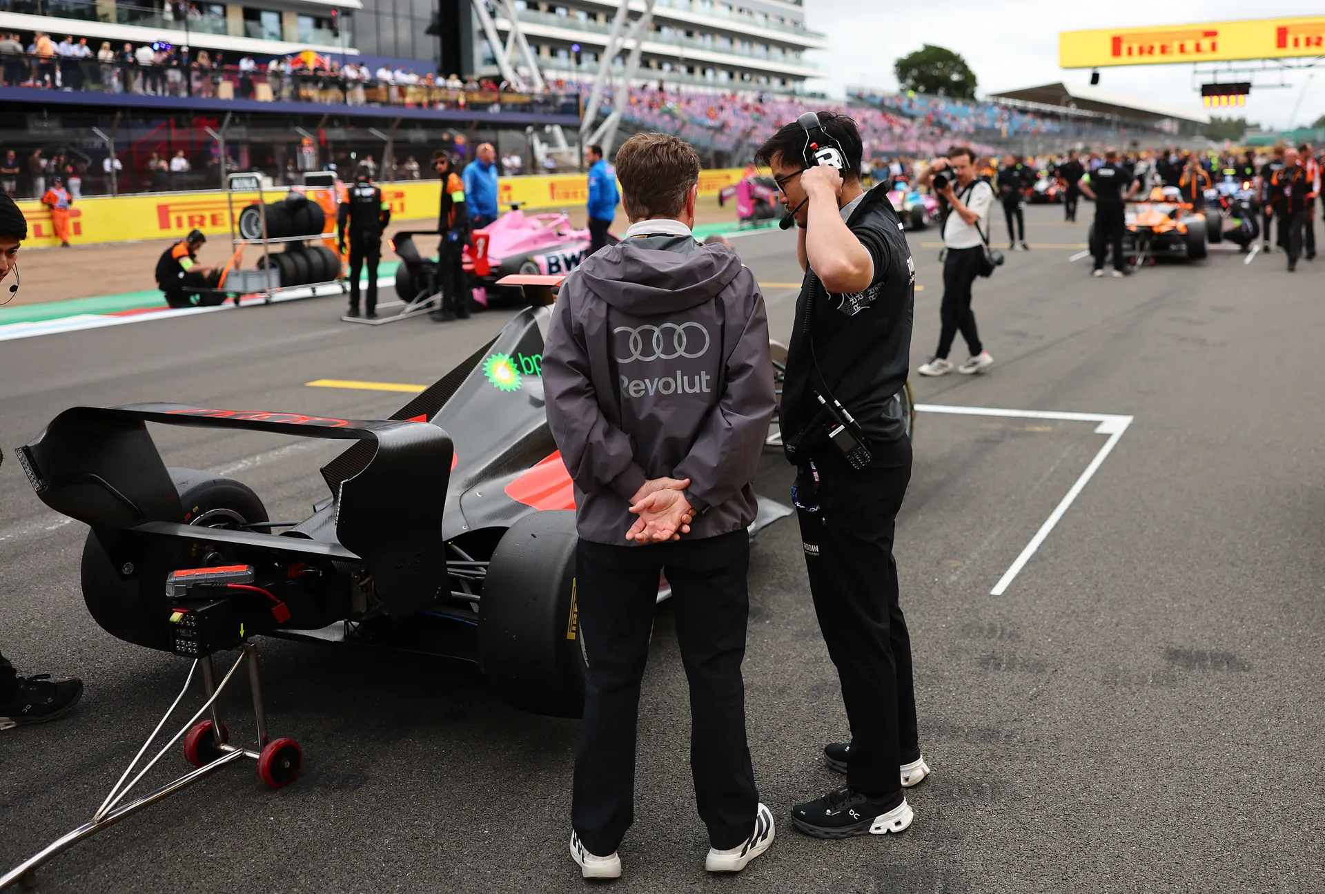 Audi Revolut F1® Team crew members and Allan McNish stand on the Silverstone starting grid beside an F1 Academy car, reviewing setup with other cars and team personnel visible along the grid and pit straight.