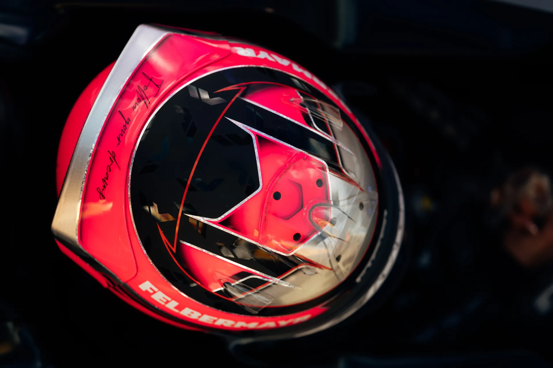 Close-up detail of a red and black racing helmet with reflective visor and signature markings, shown under paddock lighting.