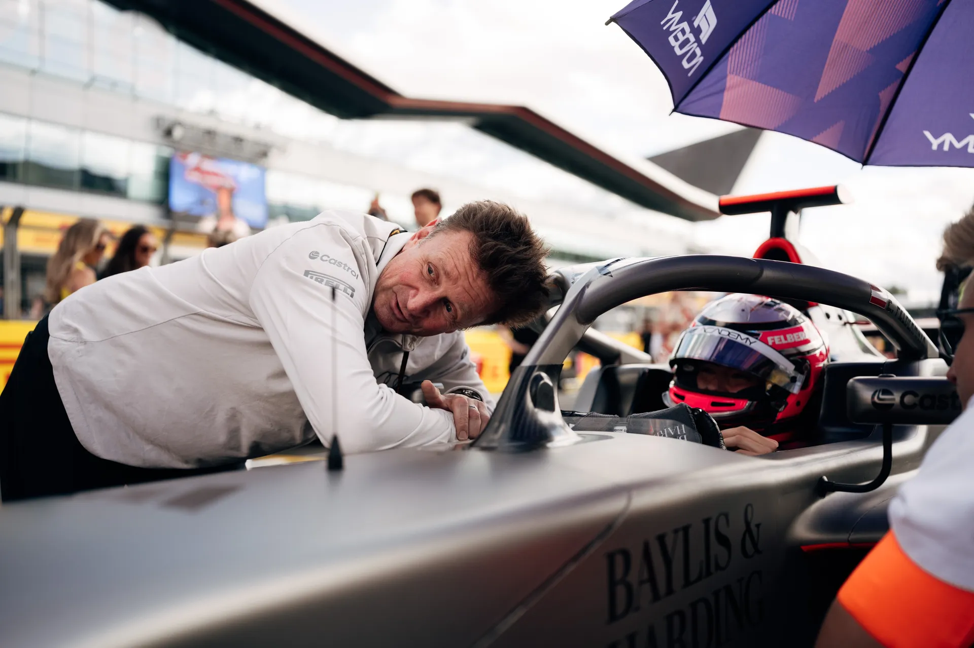 Driver in full race gear sits in the cockpit of an Audi Revolut F1® Team F1 Academy car while Allan McNish leans over the sidepod, speaking to them on the Silverstone grid.