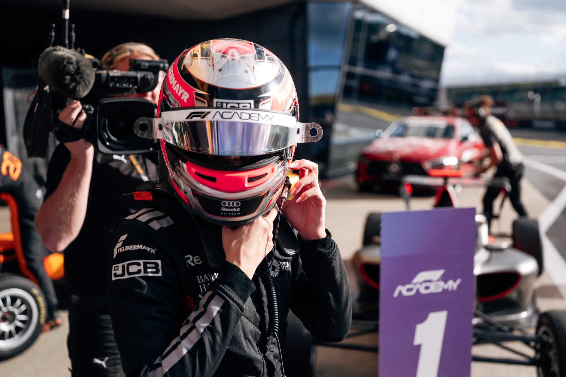 Driver wearing a full-face helmet adjusts their chin strap in the Silverstone paddock, with a camera operator filming closely and an F1 Academy P1 board visible nearby.