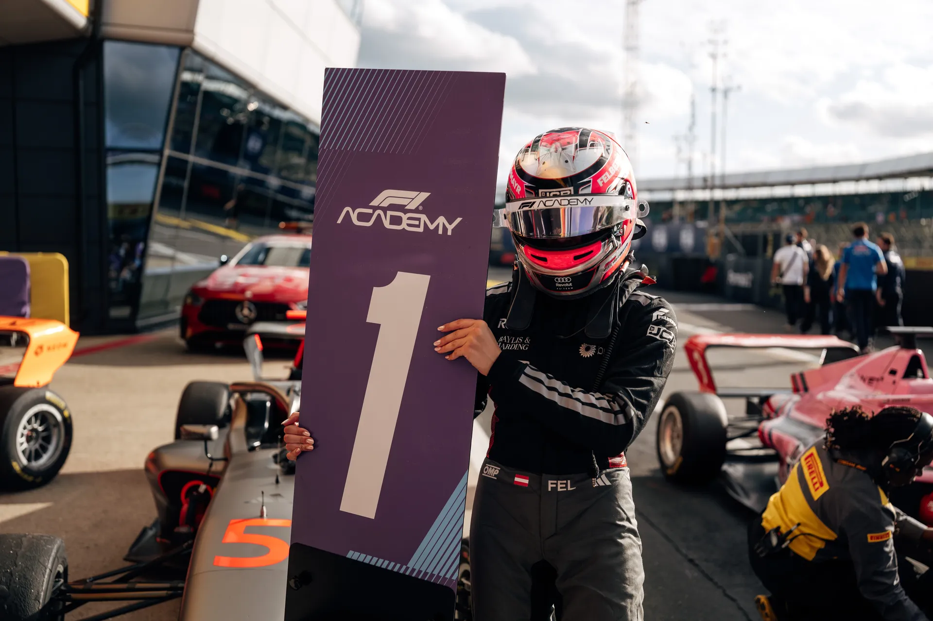 Emma Felbermayr in full race suit and helmet holds a large F1 Academy P1 position board in front of an Audi Revolut F1® Team car in the Silverstone paddock.