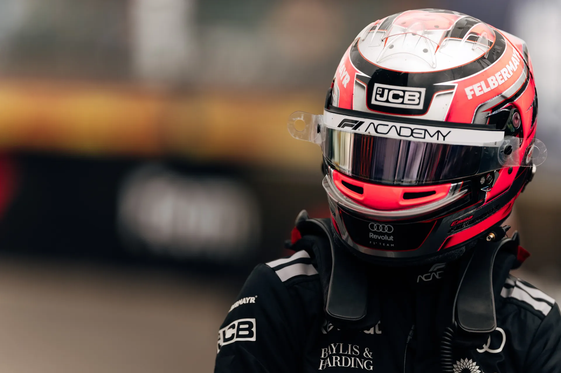 Portrait of a driver in full race suit and helmet with F1 Academy branding, standing in the paddock with blurred circuit background.