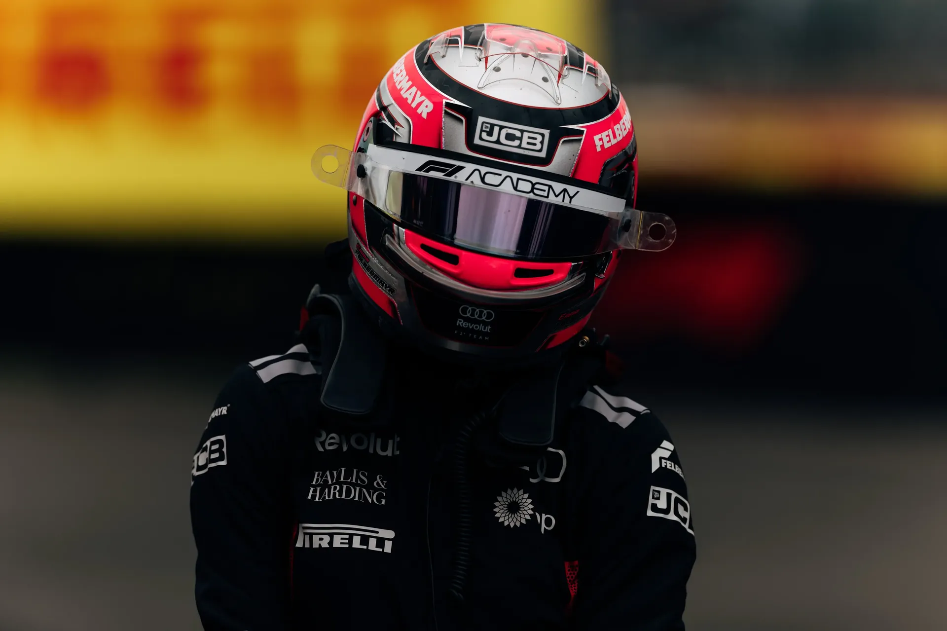 Portrait of a driver in full Audi Revolut F1® Team race gear and helmet, standing in front of a blurred Silverstone backdrop with sponsor branding visible on the suit.