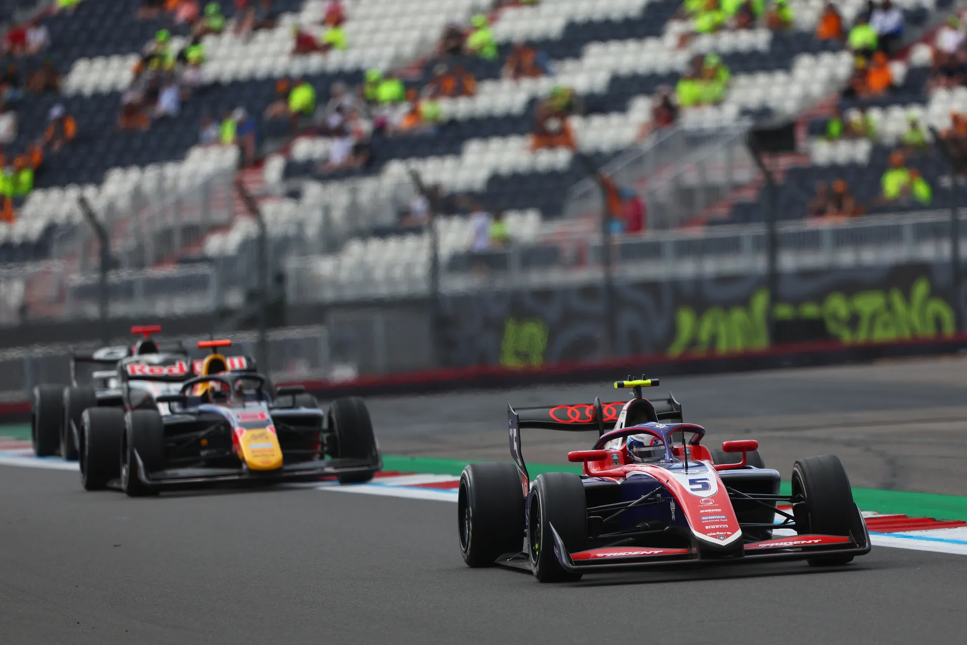 Freddie Slater drives a Trident Formula 3 car during a race at Silverstone, with another car following closely through a corner and grandstands in the background.