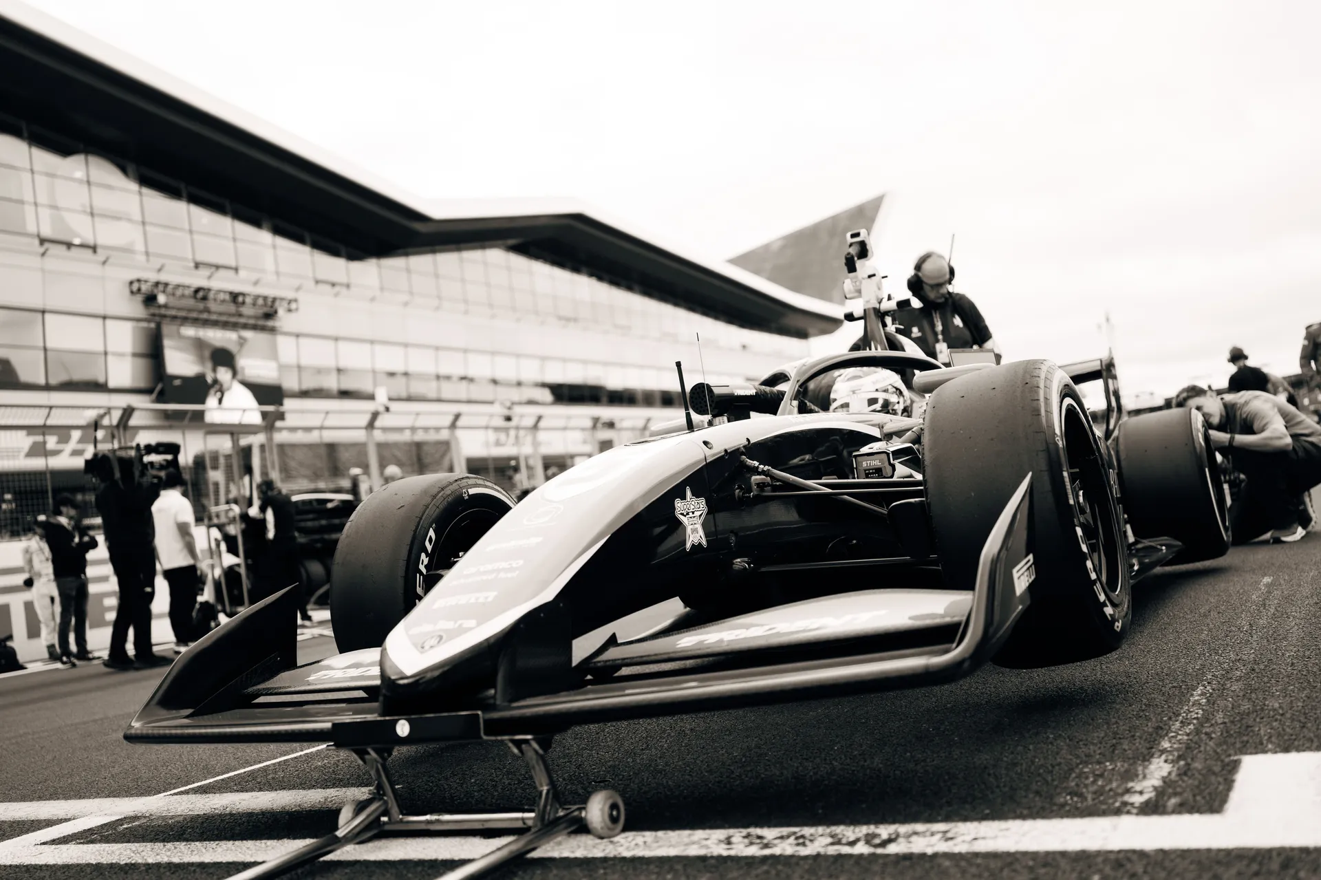 Black-and-white low-angle shot of a Trident Formula 3 car on the Silverstone grid, surrounded by team members and broadcast crew preparing the car.