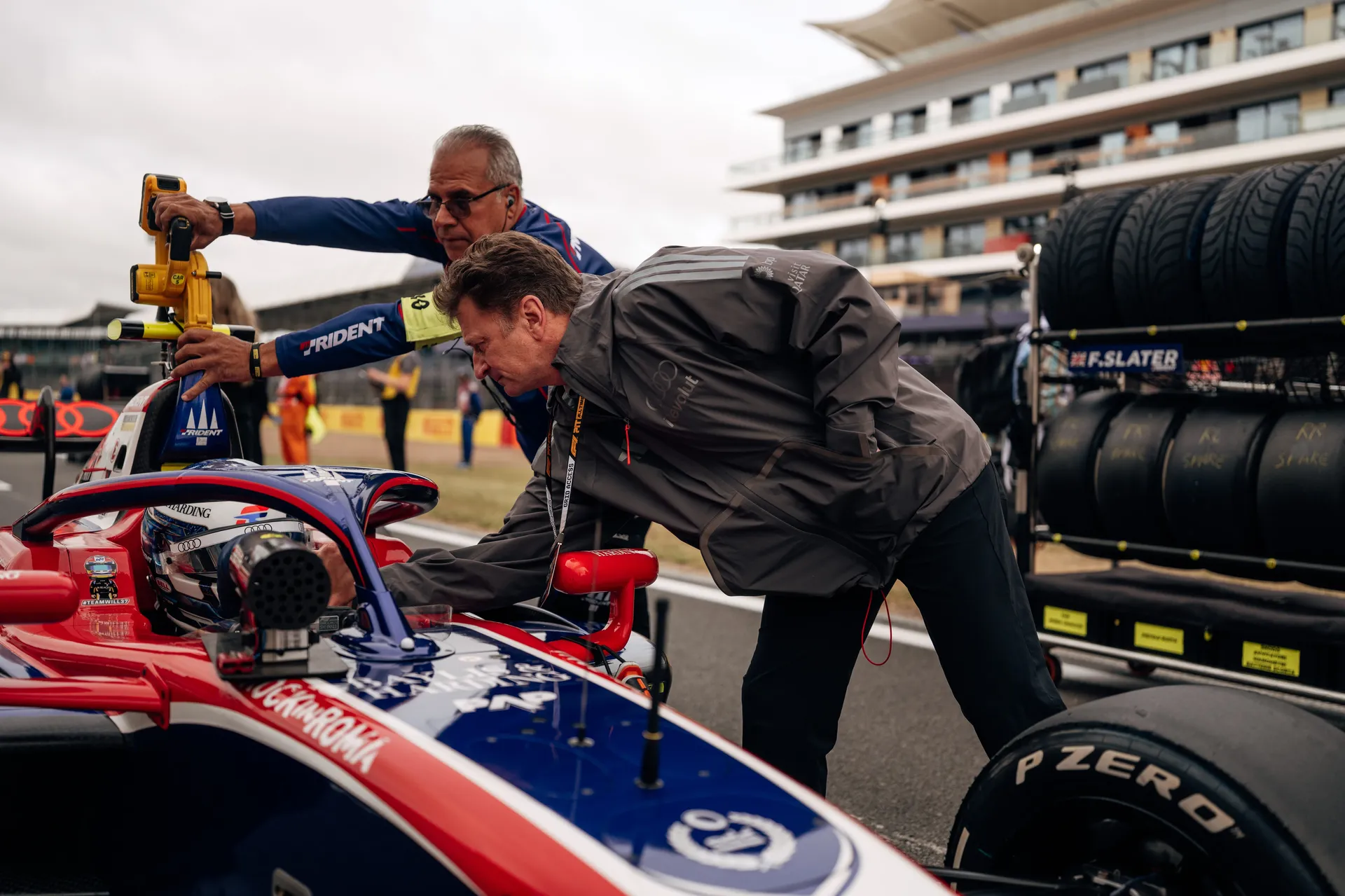 Allan McNish leans over the cockpit of Freddie Slater’s Trident Formula 3 car on the grid, speaking to the driver while a team member holds equipment beside them.