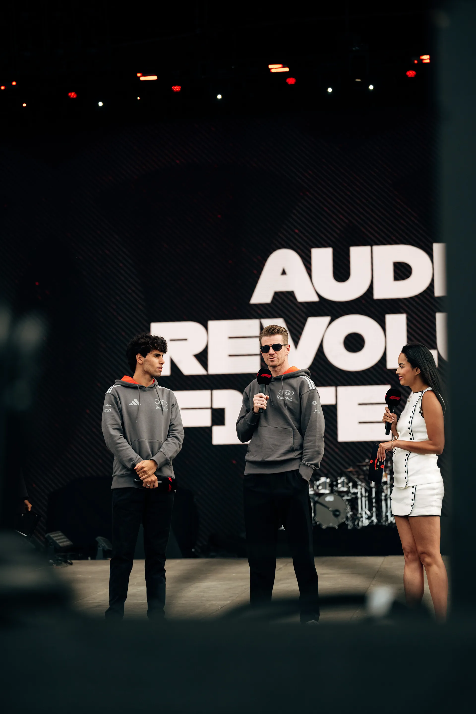 Gabriel Bortoleto and Nico Hulkenberg stand on stage with a host during the Audi Revolut F1® Team fan forum.