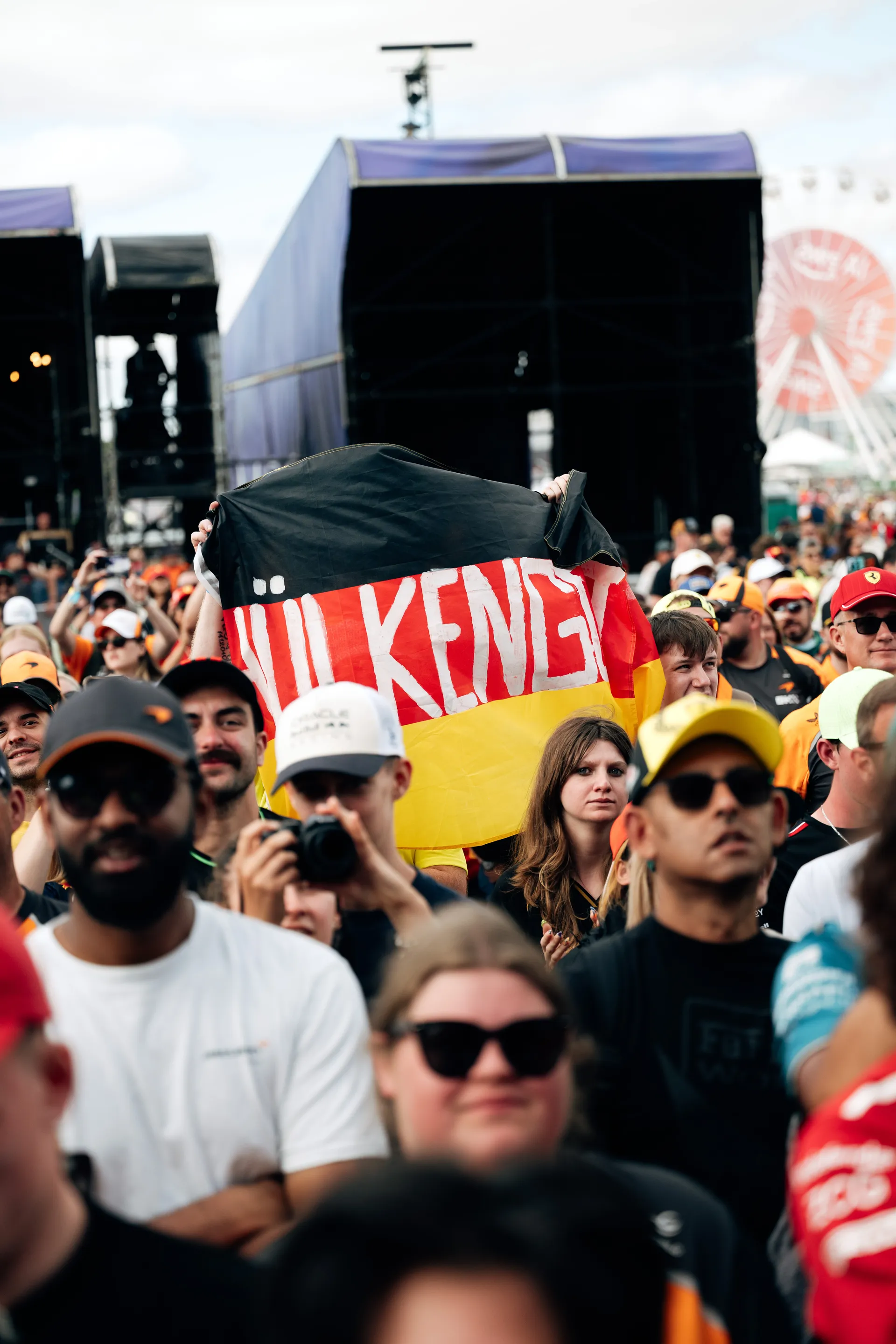 A large crowd gathers in front of the stage as a fan holds up a German flag during the Audi Revolut F1® Team fan forum.