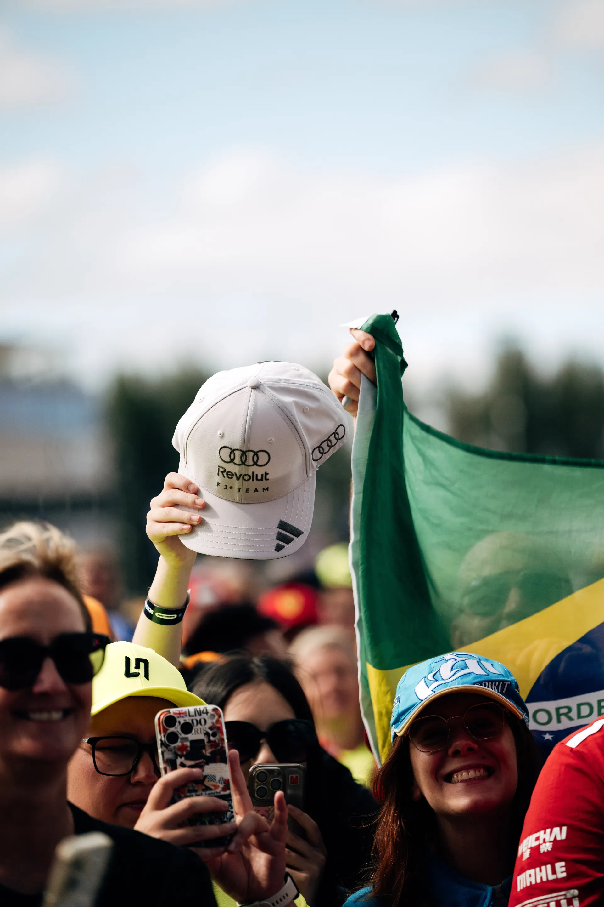 Fans in the crowd hold up a Brazilian flag and Audi Revolut F1® Team cap while taking photos at the fan forum.