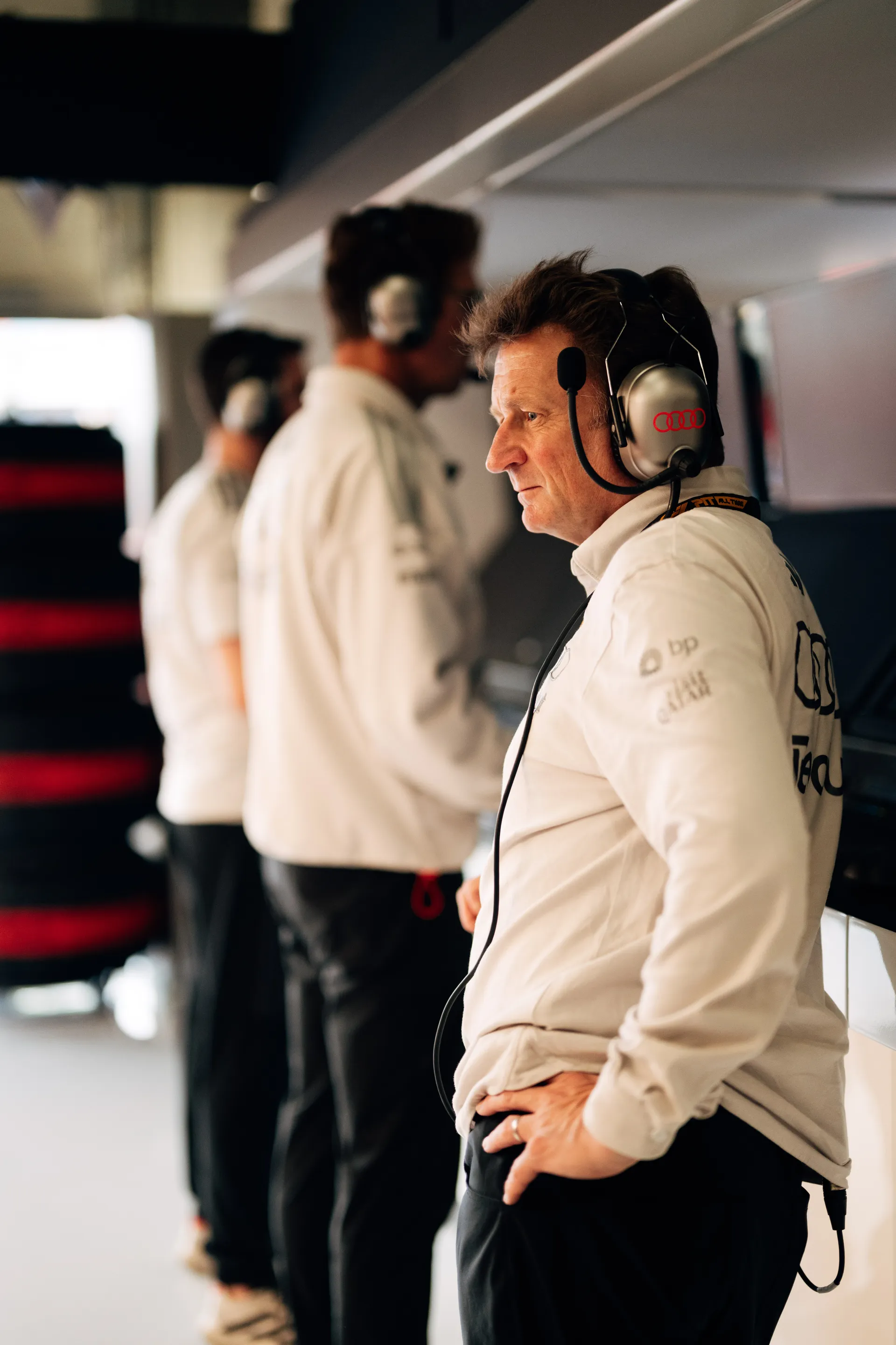 Allan McNish, Racing Director of Audi Revolut F1® Team, stands on the pit wall wearing a headset, focused on live data during Qualifying at Silverstone, with team members blurred in the background.