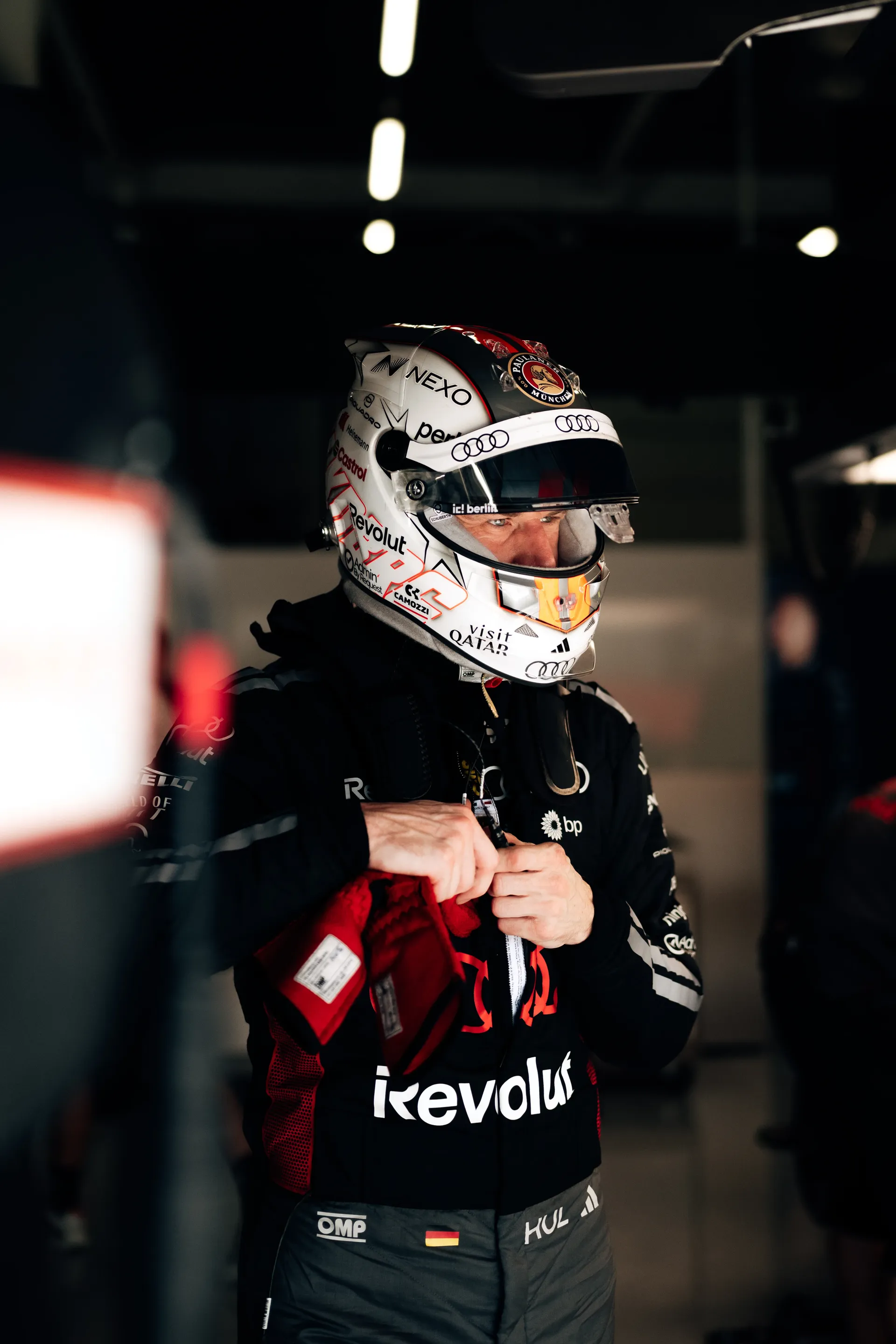 Nico Hulkenberg, wearing a white helmet with an orange visor detail, stands in the Audi Revolut F1® Team garage fastening his gloves ahead of a Qualifying run at Silverstone.