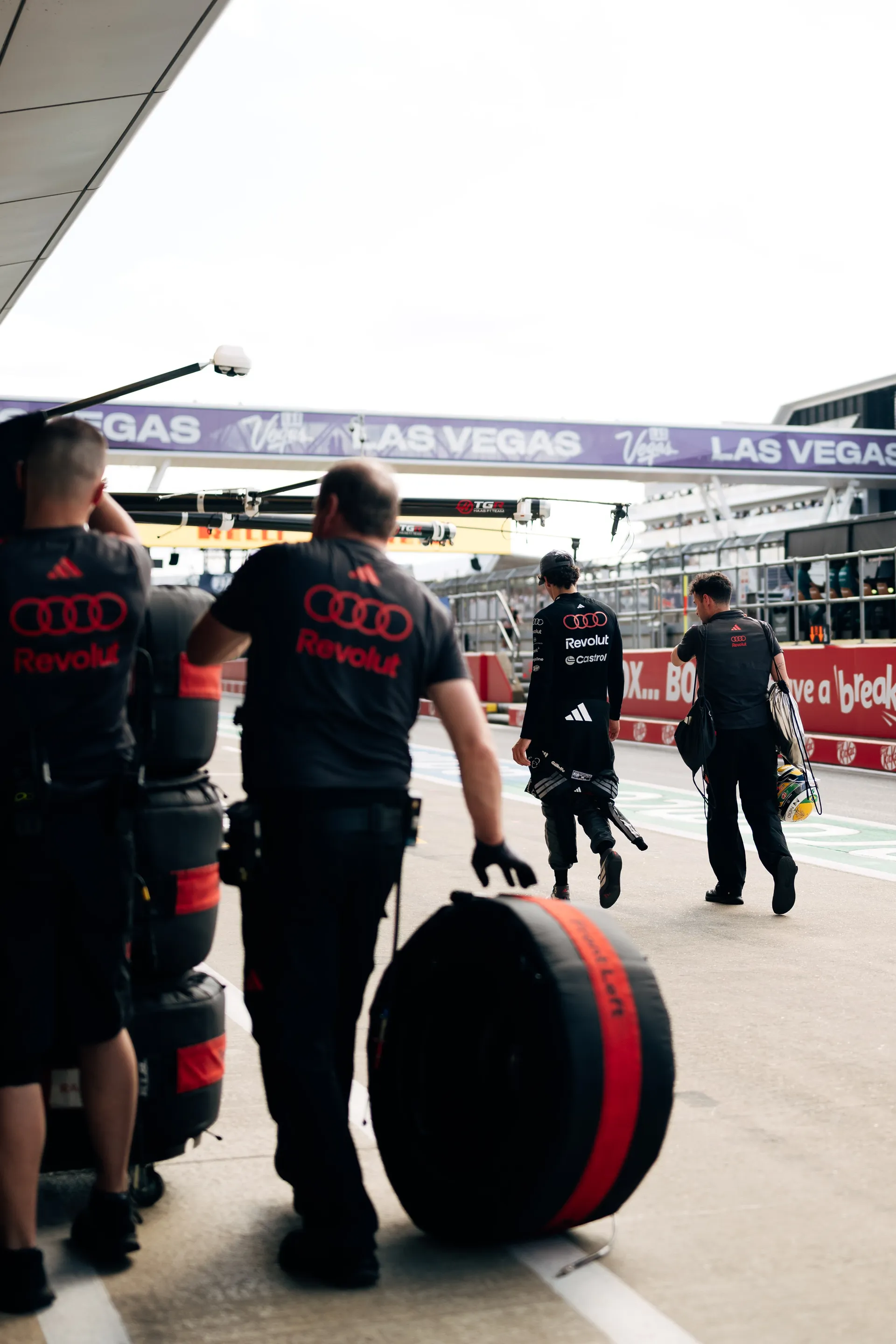 Audi Revolut F1® Team crew members transport tyres through the pit lane while a driver walks ahead carrying his helmet, heading towards the garage during the Qualifying session at Silverstone.