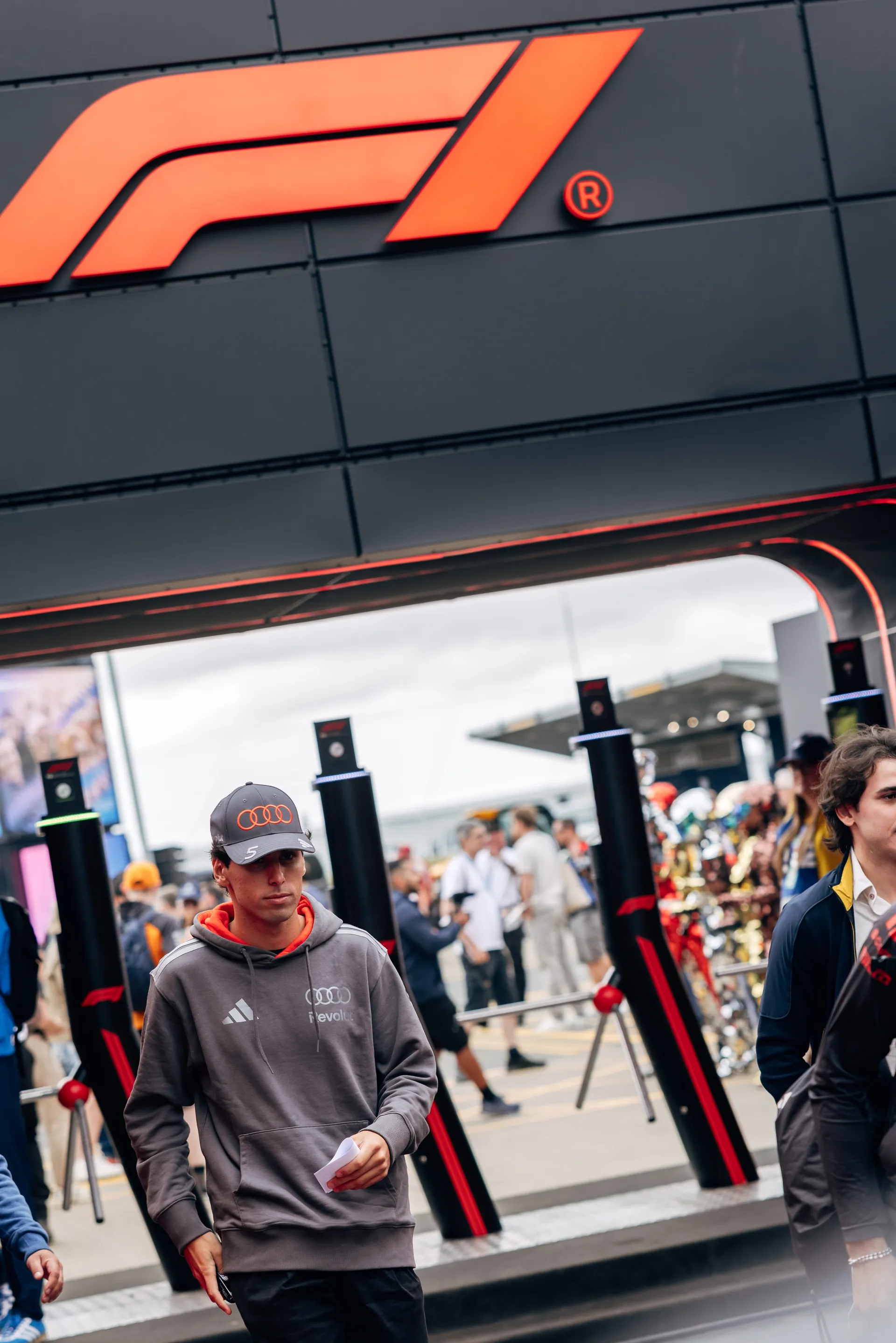 Gabriel Bortoleto walking through the paddock beneath a Formula 1 entrance structure at Silverstone, wearing Audi Revolut F1® Team kit.