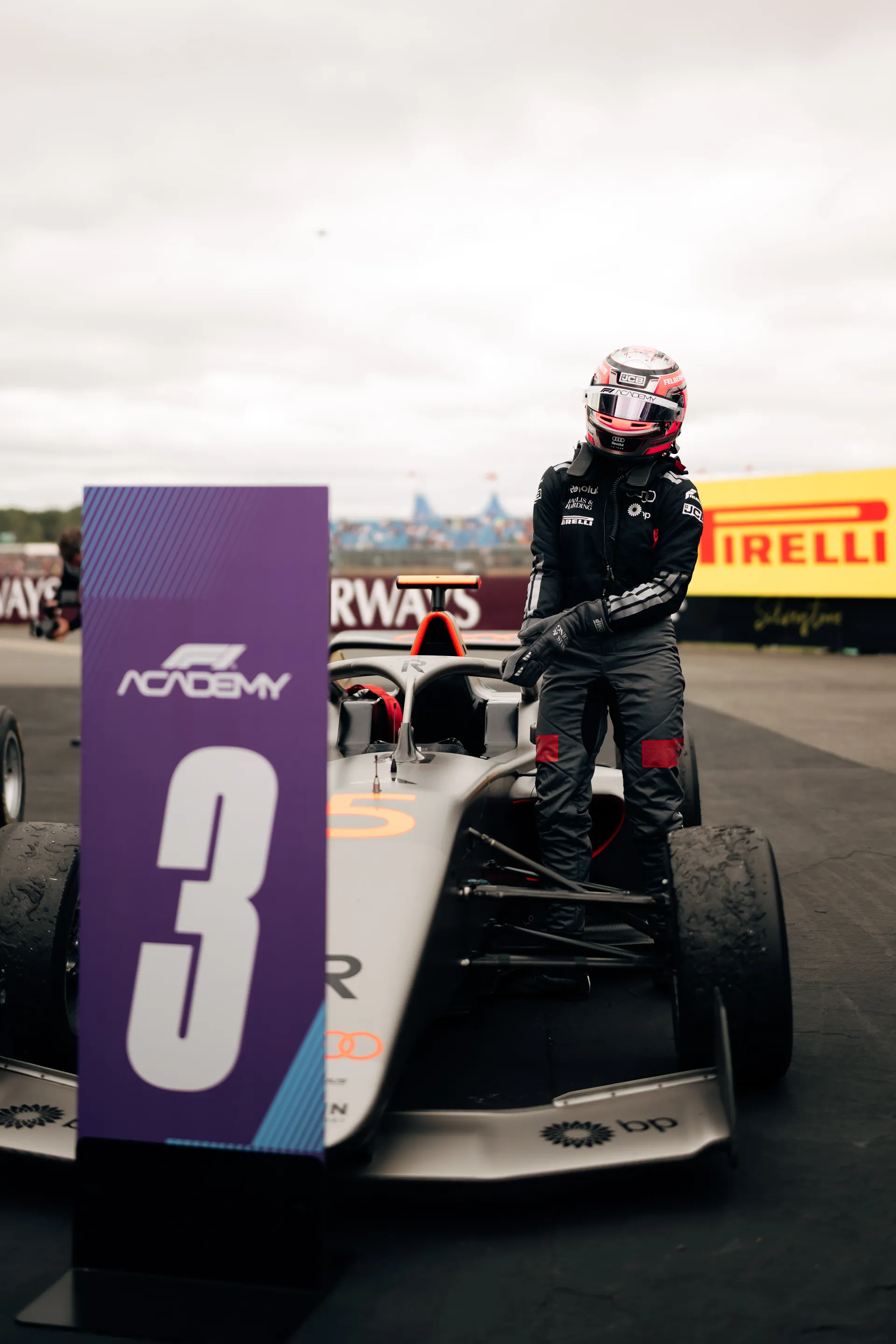 Emma Felbermayr standing beside her car in full race suit and pink helmet near an F1 Academy grid marker.