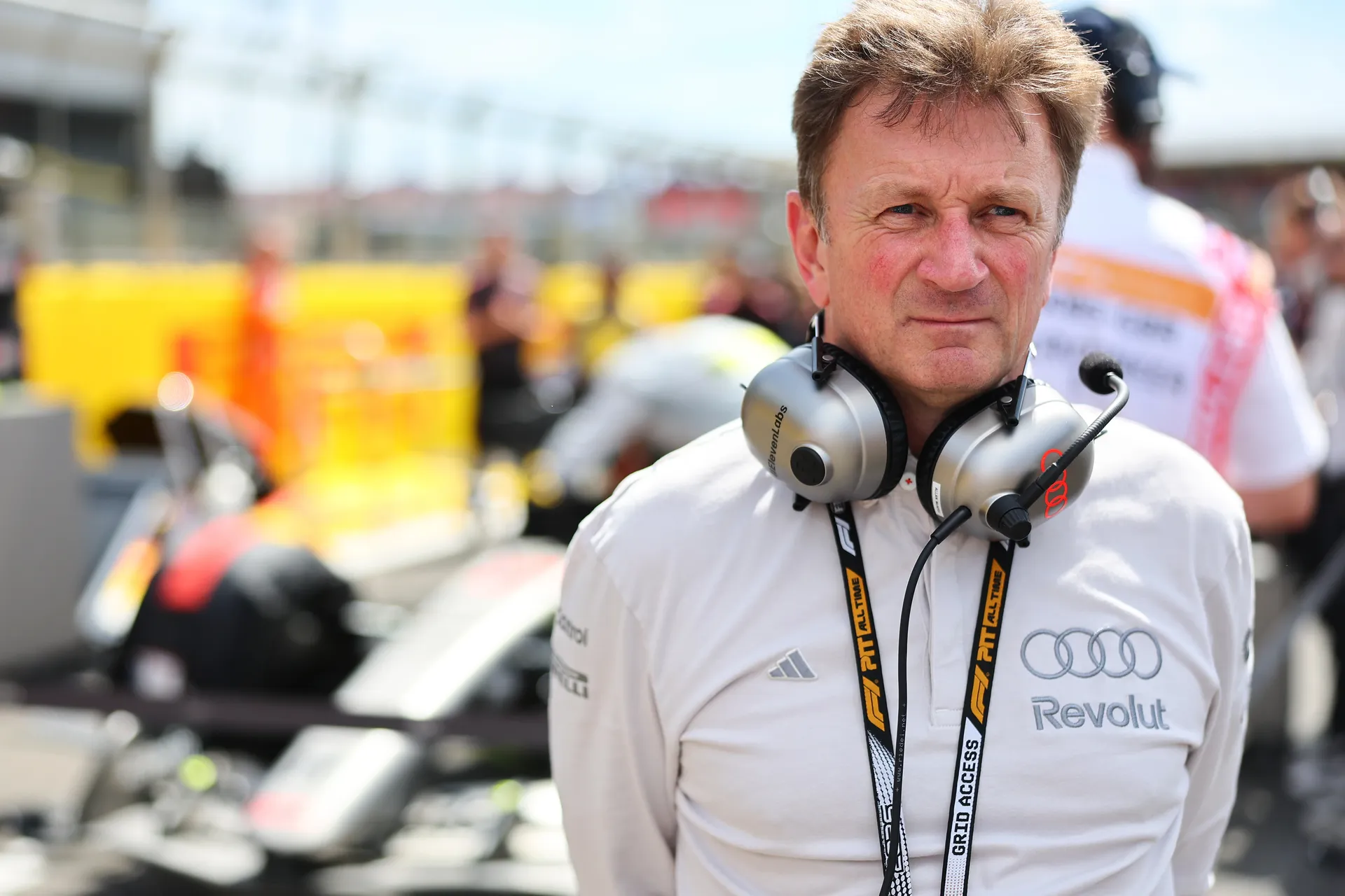 Audi Revolut F1® Team Allan McNish standing on the grid with headset and lanyard, with an Audi R26 in the background.