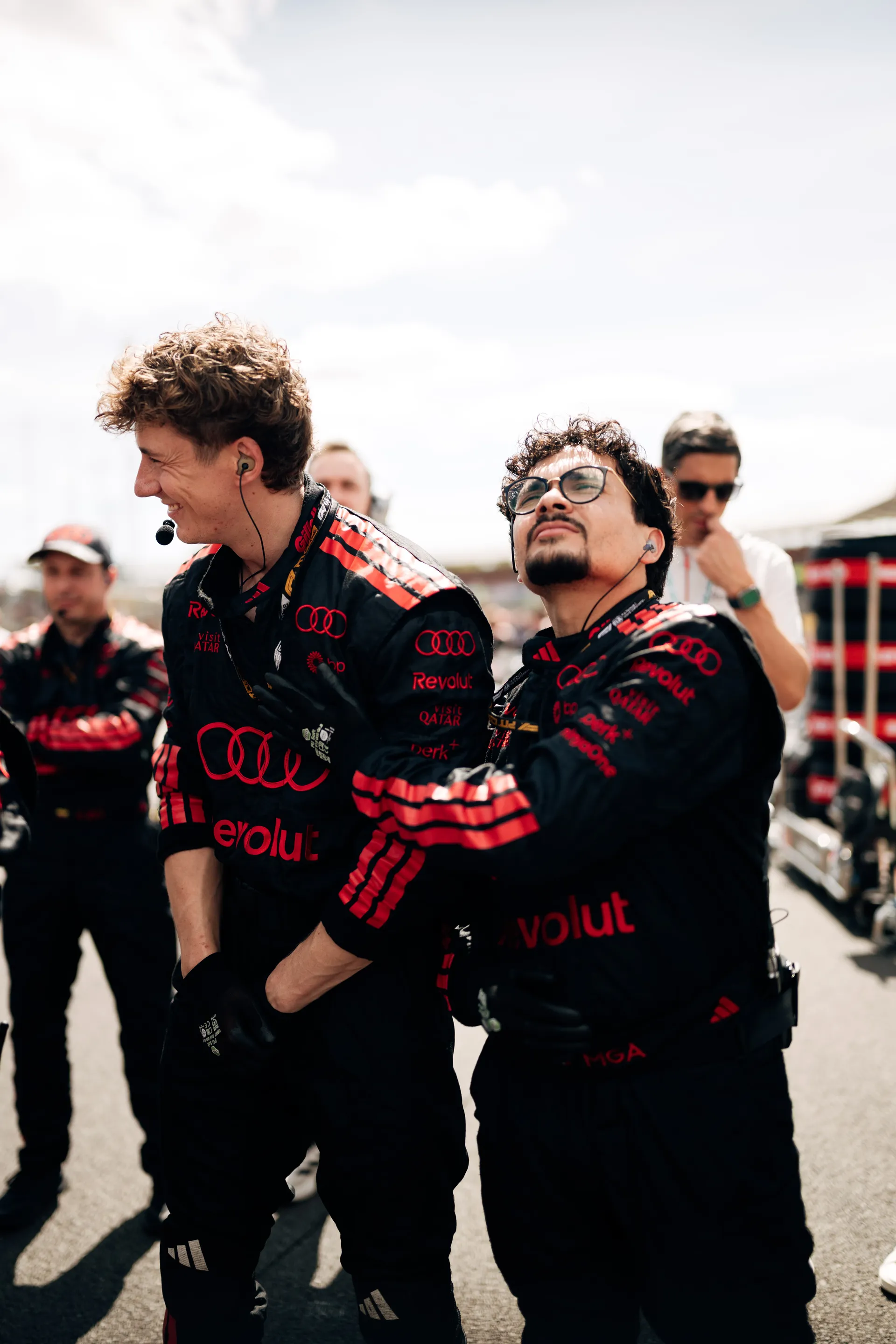 Two Audi Revolut F1® Team crew members in black race suits stand on the grid, wearing headsets and looking upwards, focused on pre-race preparations.