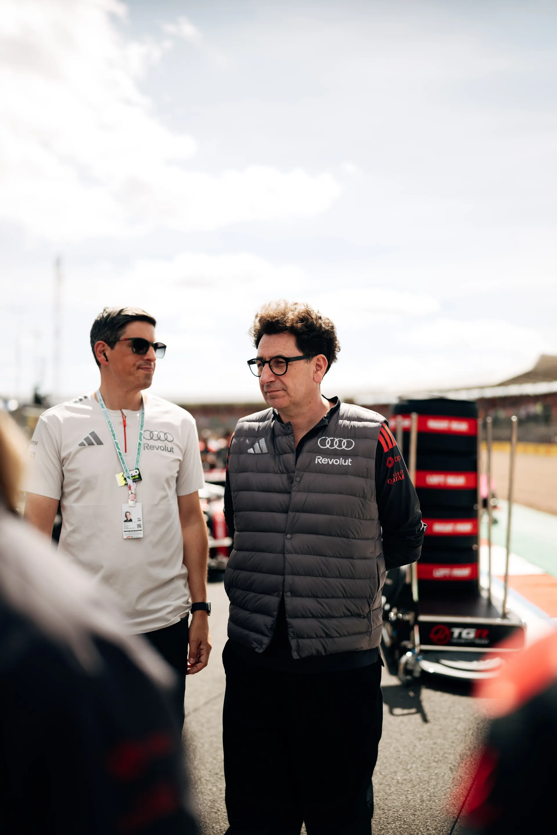 Mattia Binotto stands trackside with an Audi Revolut F1® Team colleague, both wearing team gear and sunglasses, observing activity in the pit lane.