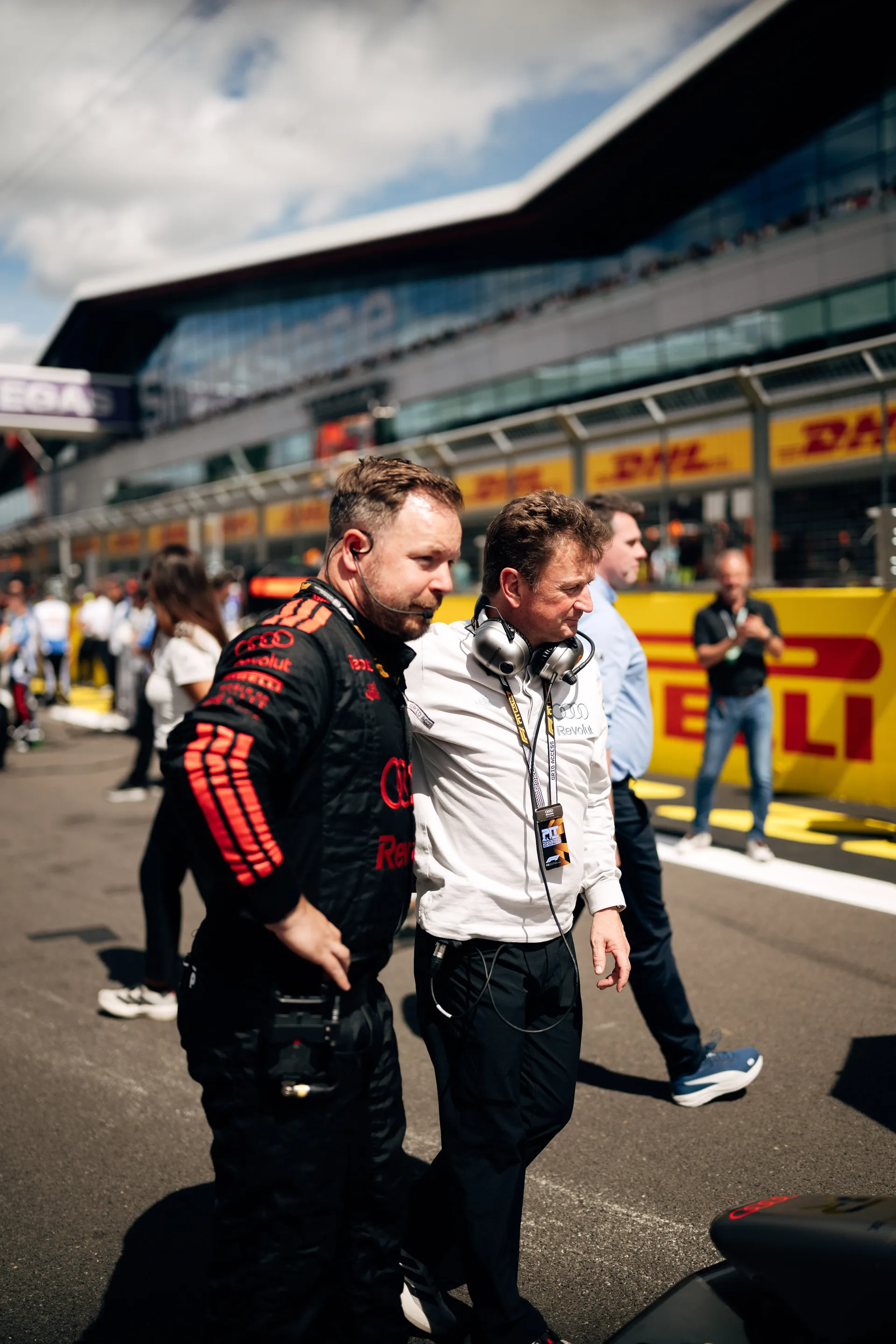 Two Audi Revolut F1® Team crew members walk along the pit lane in conversation, wearing headsets during race preparations.