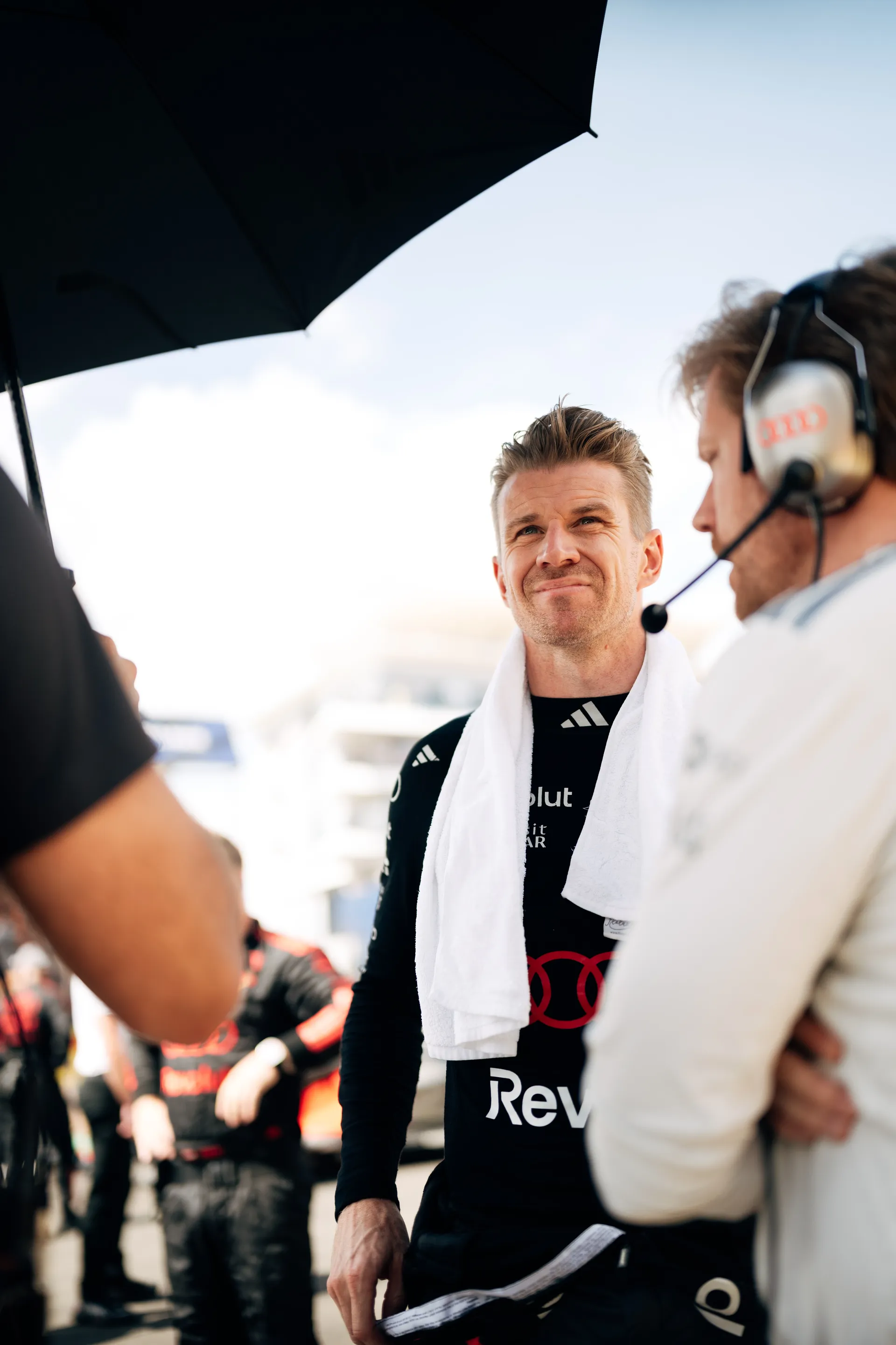 Nico Hulkenberg stands under an umbrella in the garage area wearing his race suit and a towel, listening to a team member through a headset.