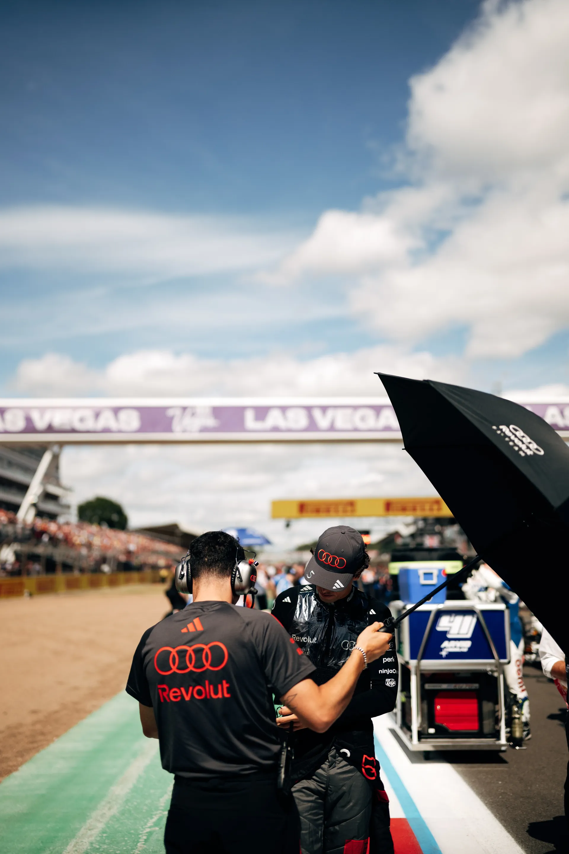 A team member assists Gabriel Bortoleto on the grid under an umbrella, adjusting his gear before the session.