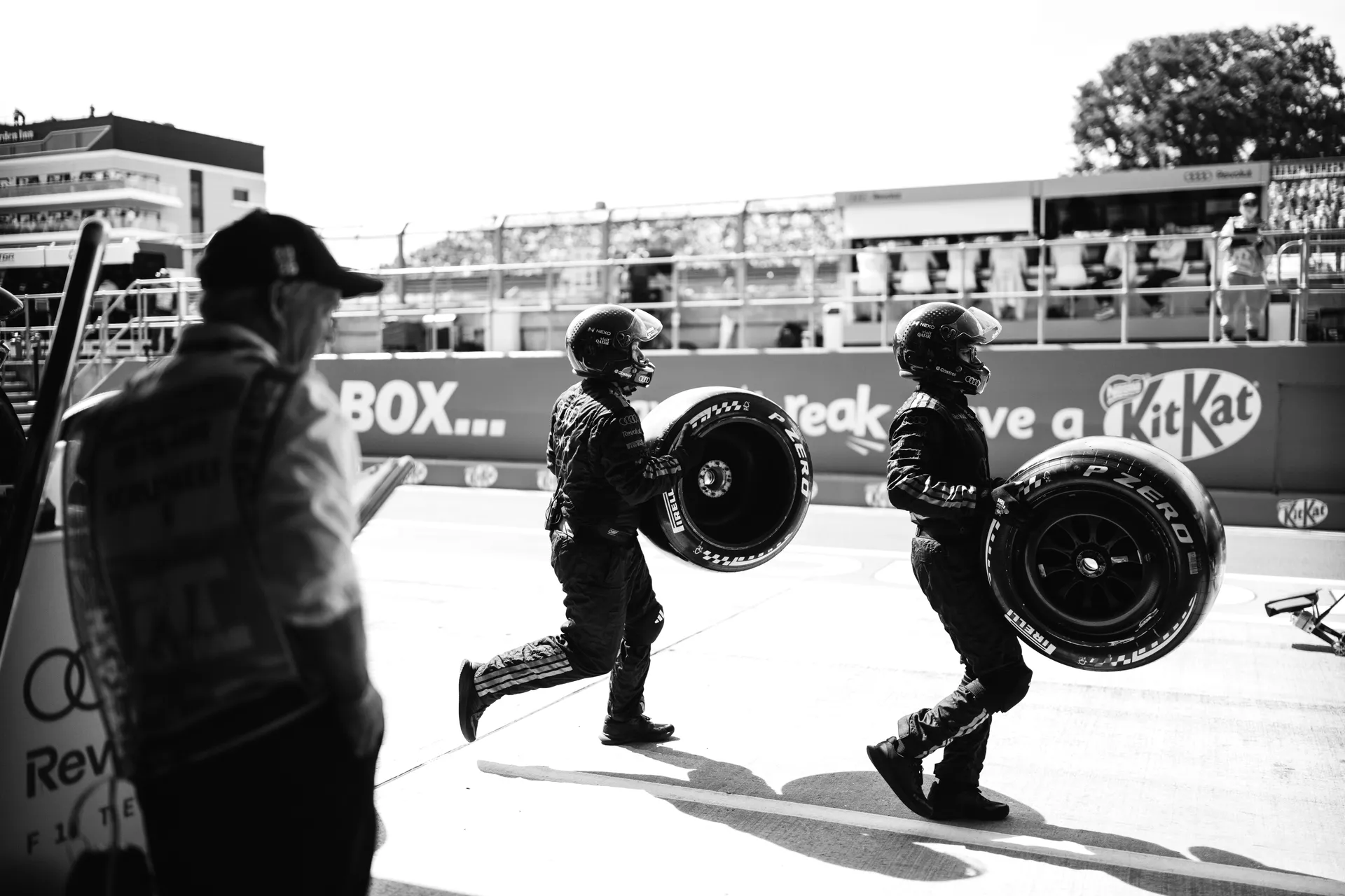Two Audi Revolut F1® Team pit crew members carry tyres across the pit lane during a pit stop sequence in black and white.