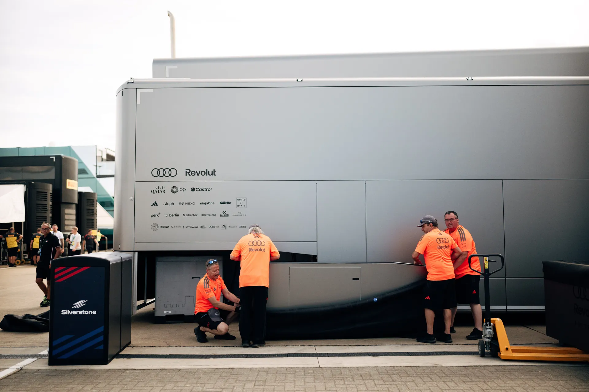 Team staff in orange shirts work behind the Audi Revolut F1® Team garage unit at Silverstone, handling equipment and logistics.