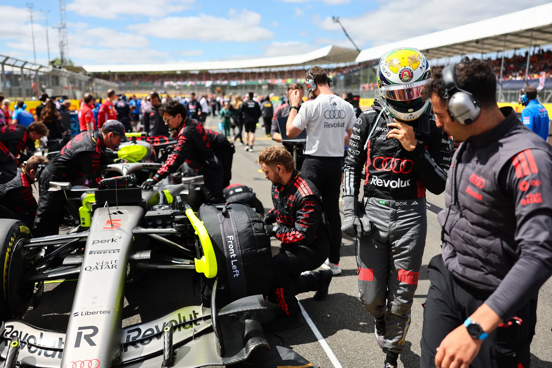 Gabriel Bortoleto adjusting his earpiece on the grid while Audi Revolut F1® Team mechanics work on the car beside him.