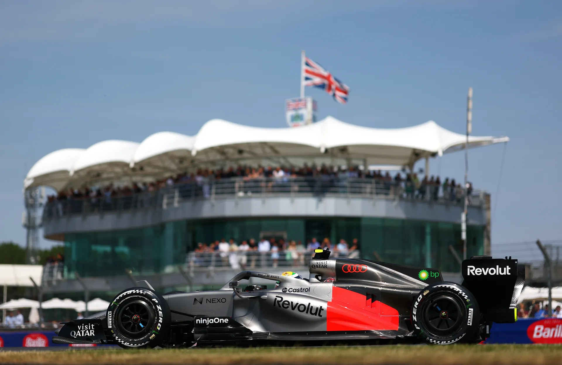 The Audi R26 runs on track at Silverstone with grandstands and a Union Jack flag in the background.