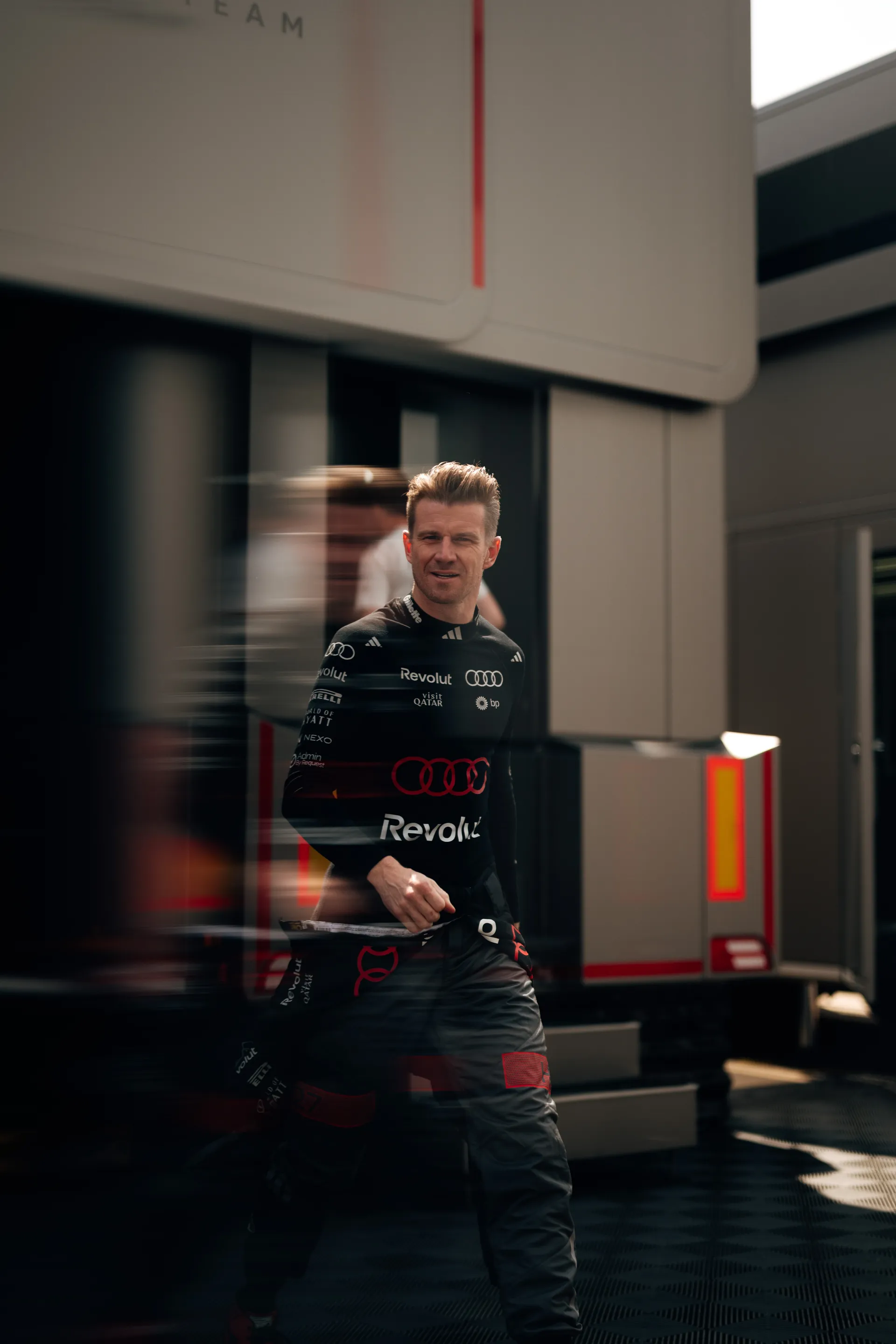 Nico Hulkenberg walks past the Audi Revolut F1® Team garage with blurred movement crossing the frame.