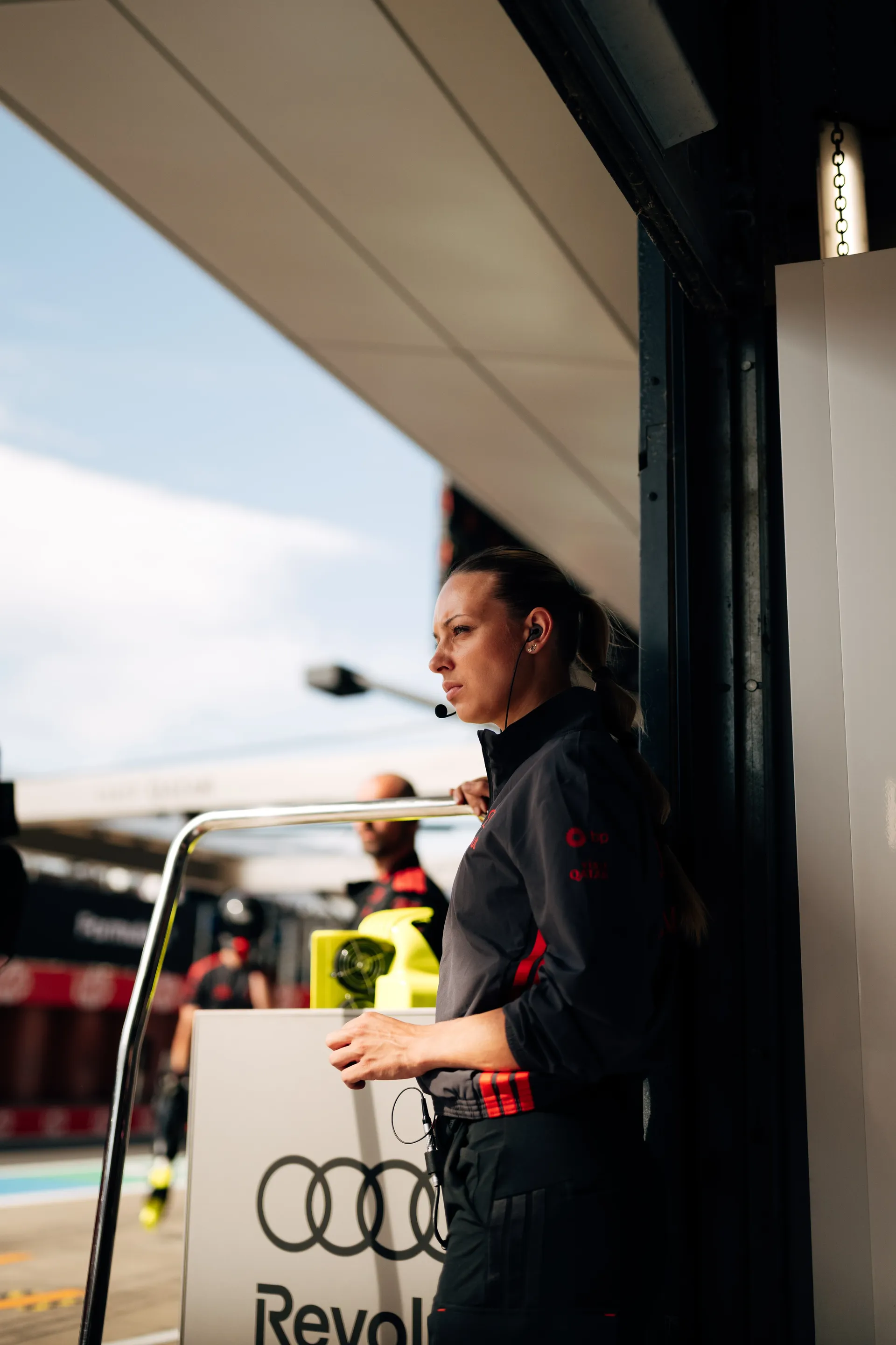 A female Audi Revolut F1® Team crew member stands at the pit wall wearing a headset and observing the track.