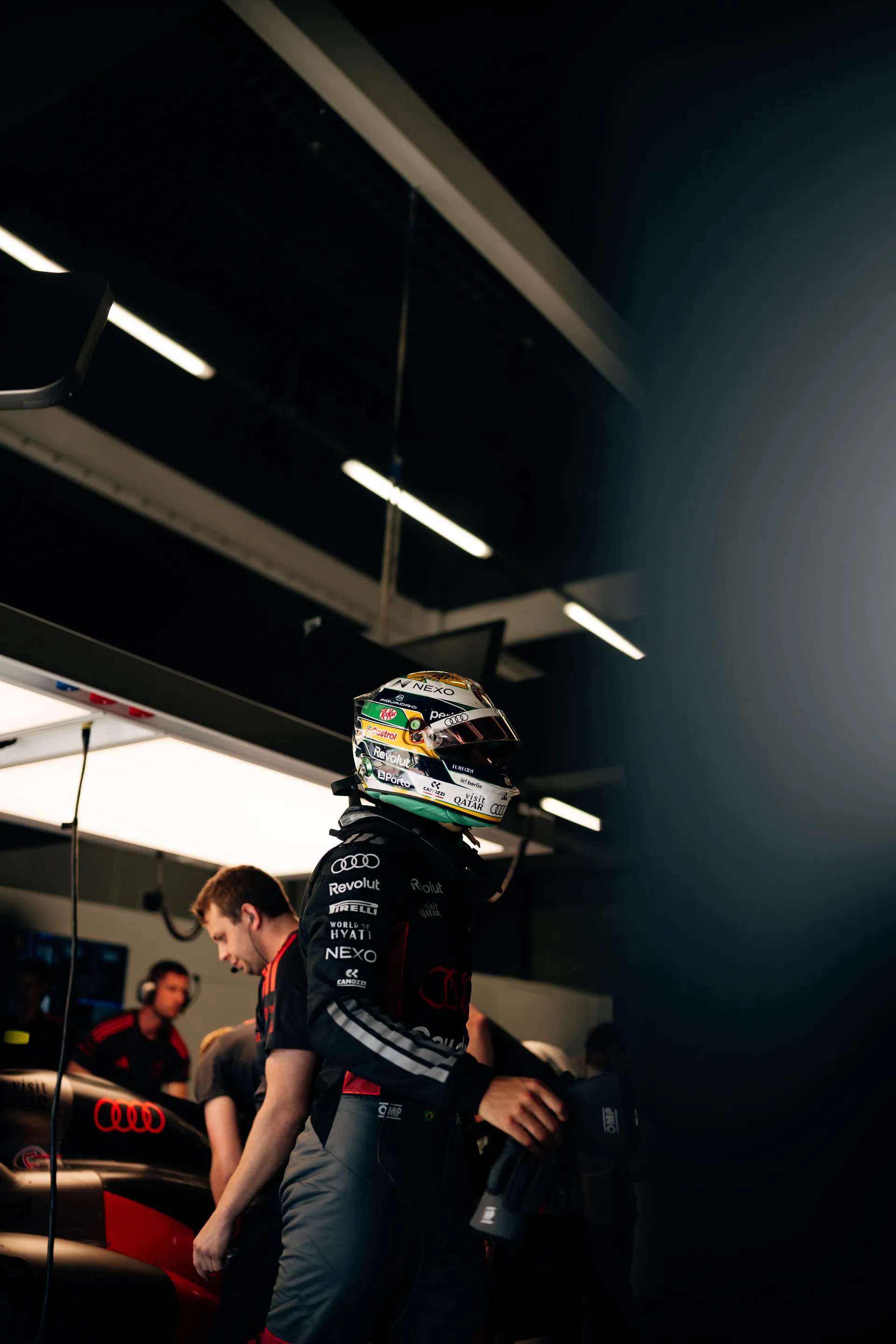 Gabriel Bortoleto stands in full race gear inside the Audi Revolut F1® Team garage while crew members prepare the Audi R26 around him.