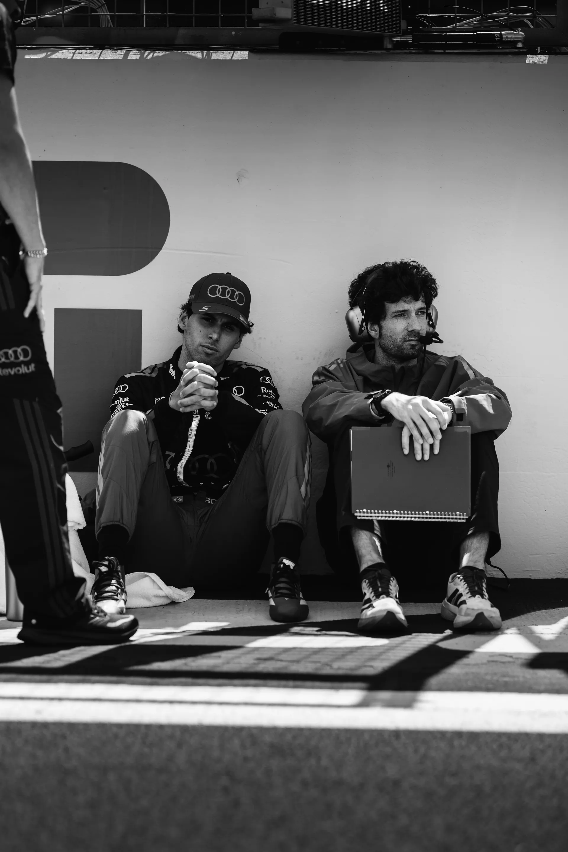 Gabriel Bortoleto sits on the ground beside a team member, focused and waiting during pre-race preparations at Silverstone.